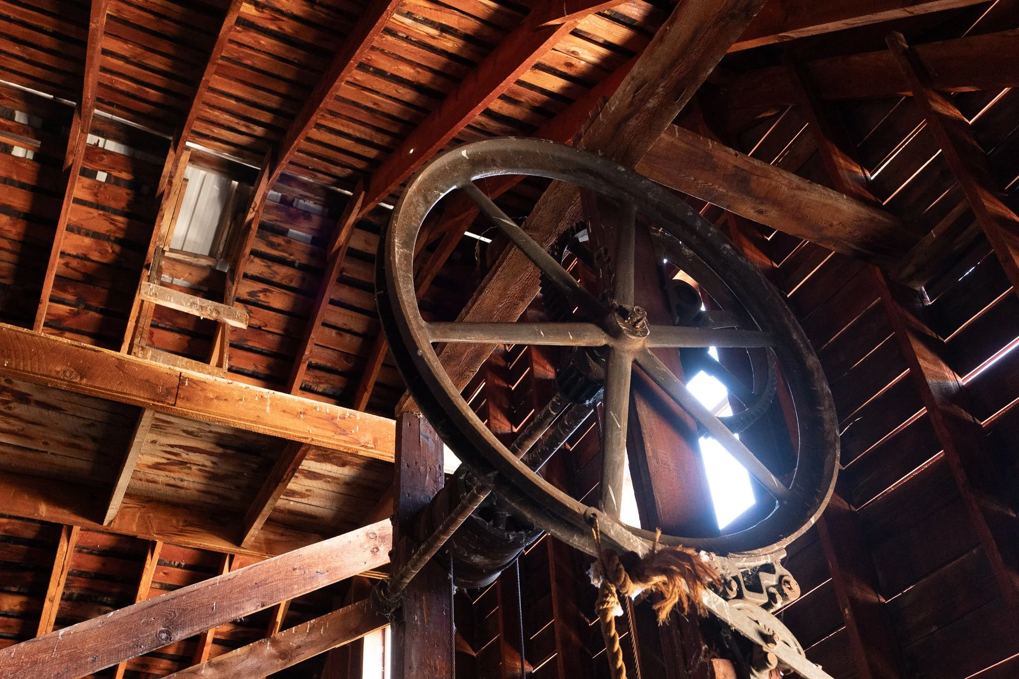 Large weathered wooden pulley wheel mounted among exposed timber beams and roof rafters inside an old ranch barn.