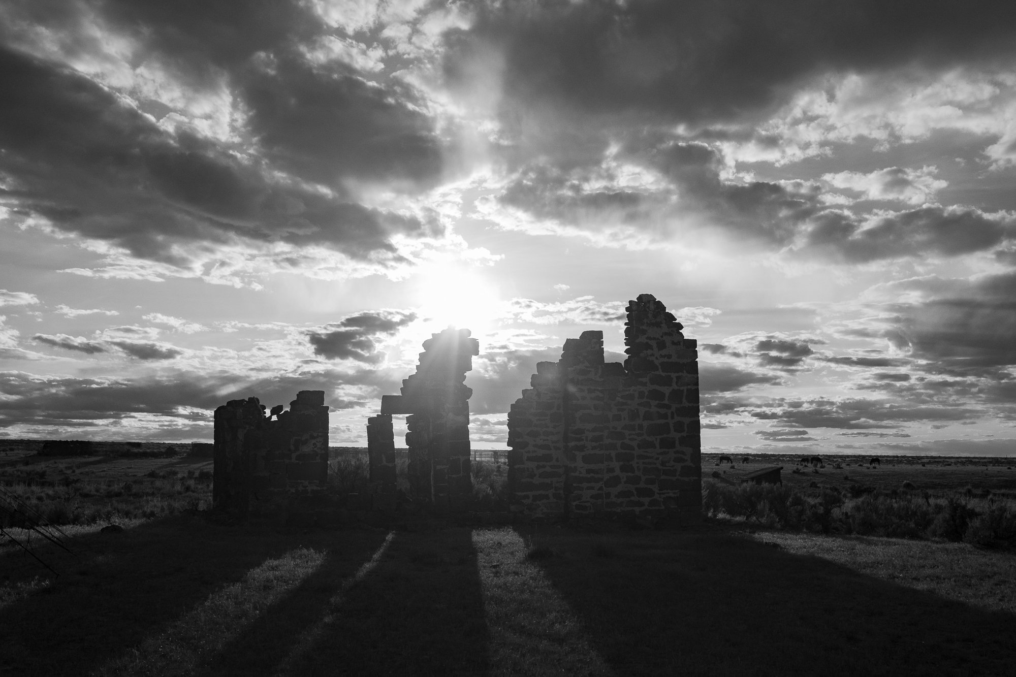 Black-and-white photograph of crumbling stone ranch building walls silhouetted against a bright, cloud-filled sky over open Western grassland.