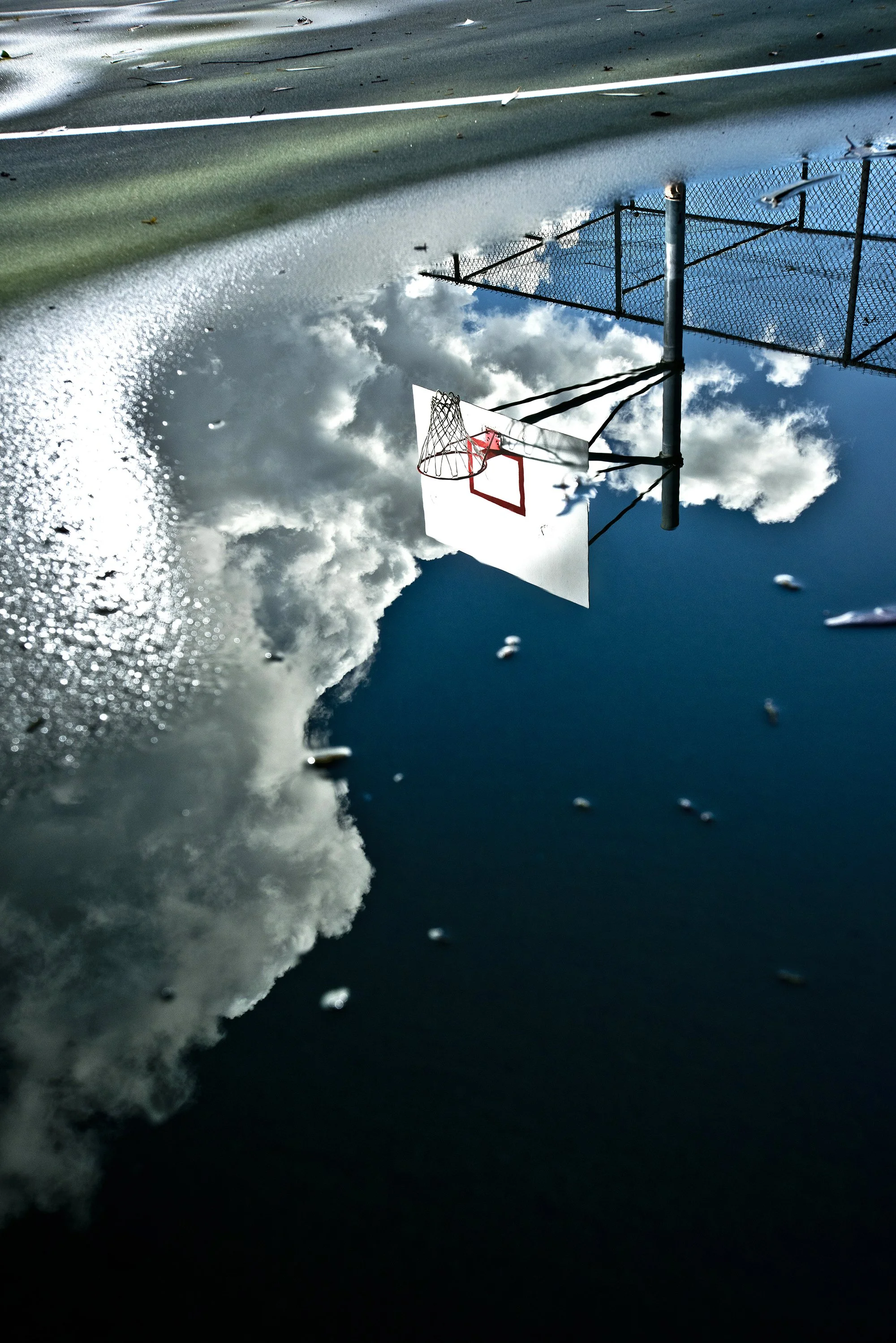Basketball hoop and backboard reflected in water beneath a partly cloudy sky.