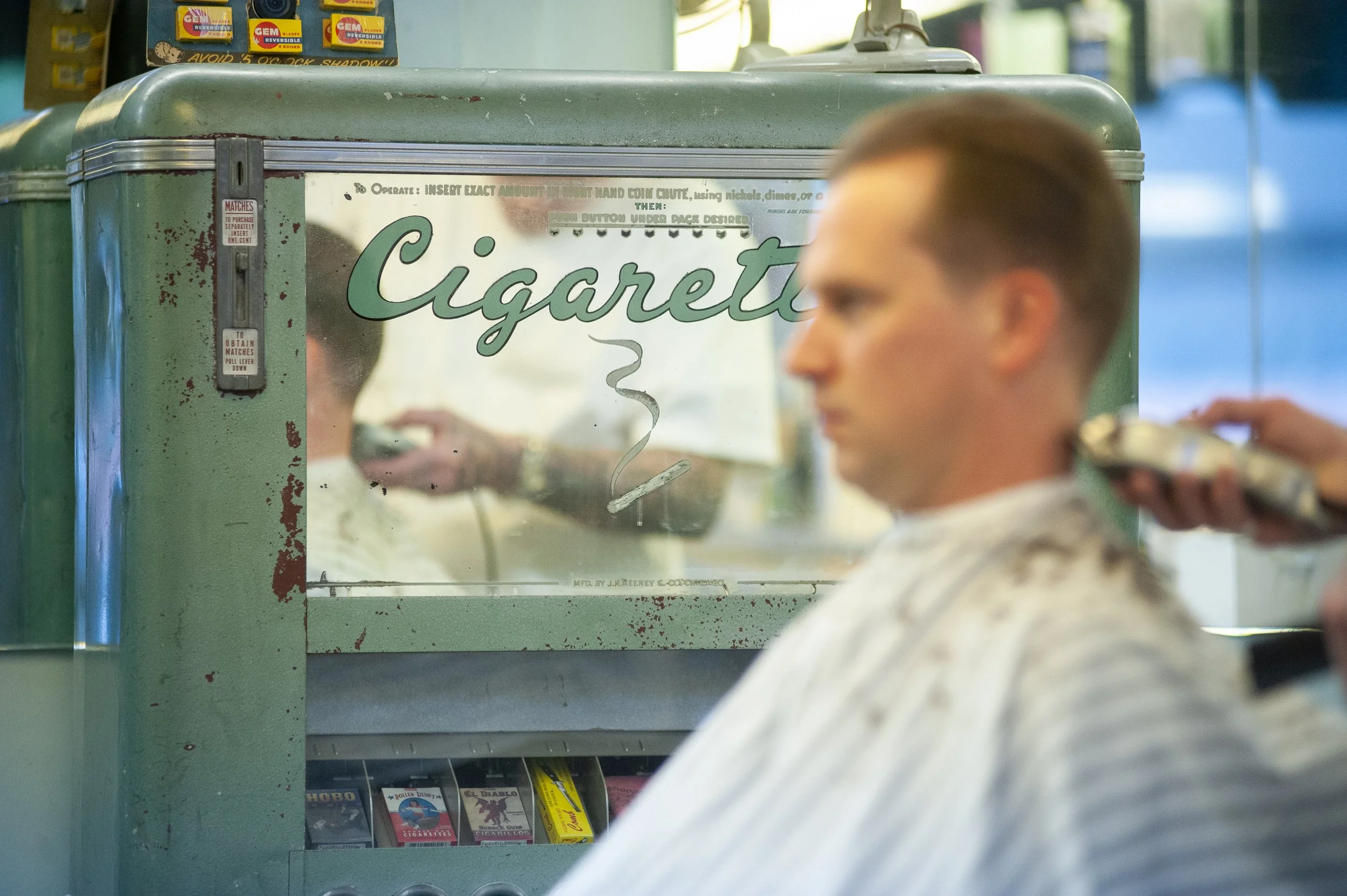Vintage green cigarette vending machine at Sweeney Todd's Barbershop in Los Angeles, with a caped client being clippered in the foreground