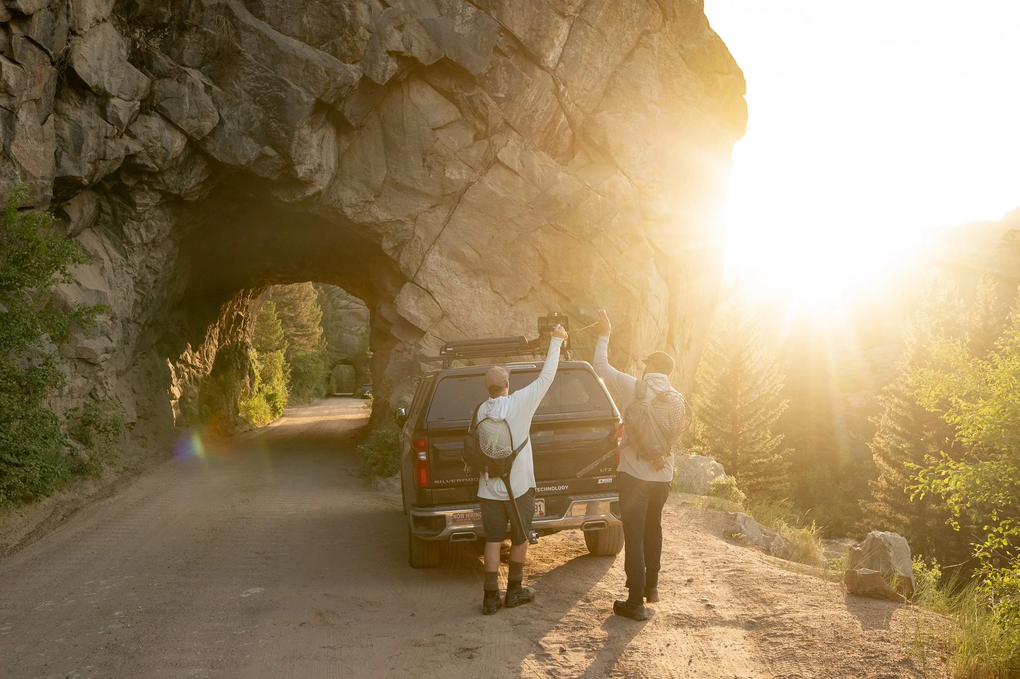 Two fly anglers stand beside a truck near a rock tunnel in warm sunlight