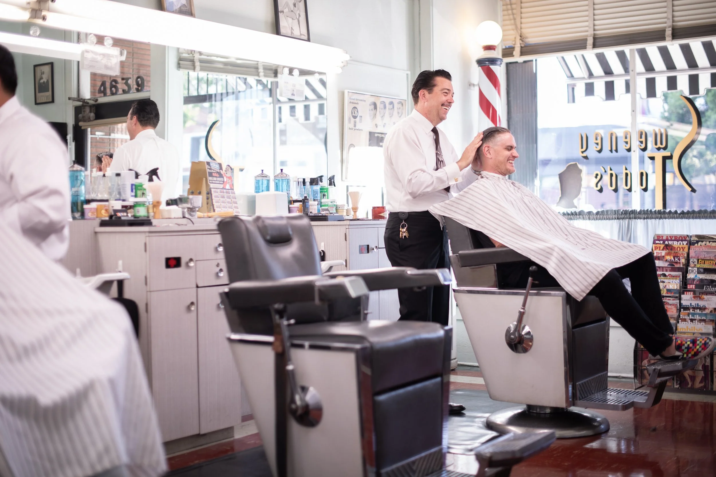 Wide interior shot of Sweeney Todd's Barbershop with barber adjusting a caped client's cape in a vintage chair, barber pole and gold window sign visible in the background