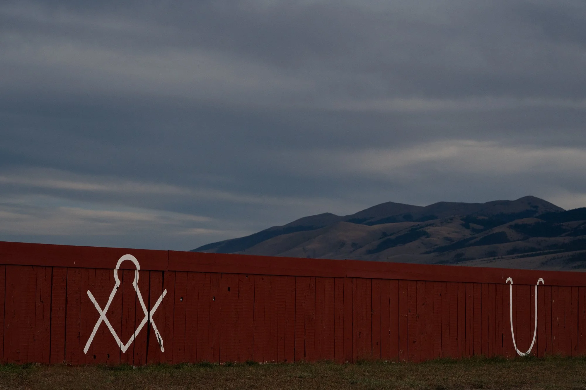 Red wooden ranch fence marked with white livestock brands, with dark mountain ridges and an overcast sky in the background.