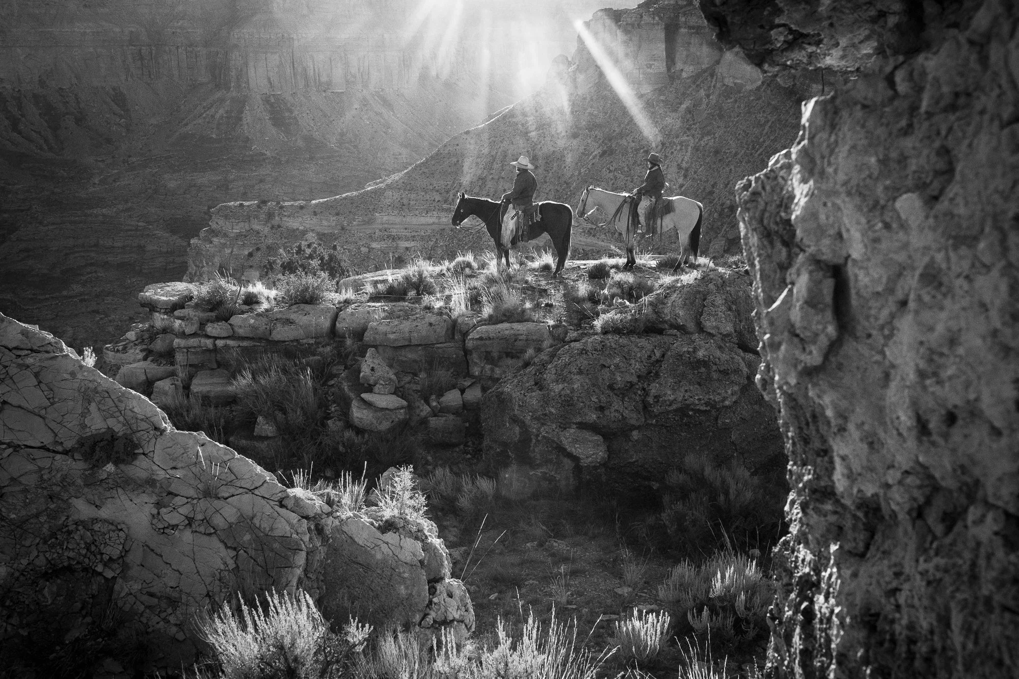 Two cowboys on horseback silhouetted against dramatic light rays breaking through canyon walls in black and white