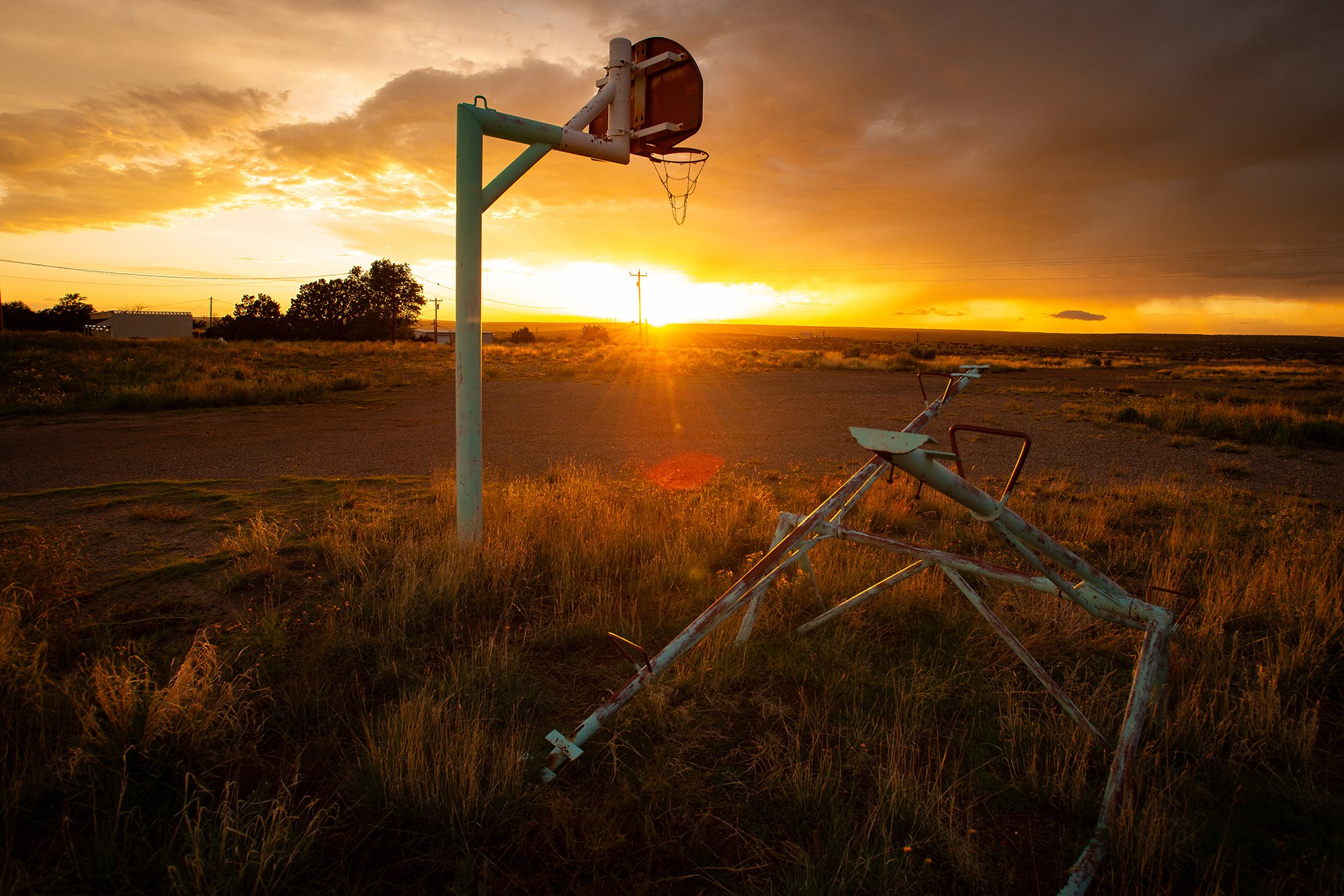 Basketball hoop standing in a rural field at sunset beside an overturned piece of farm equipment.