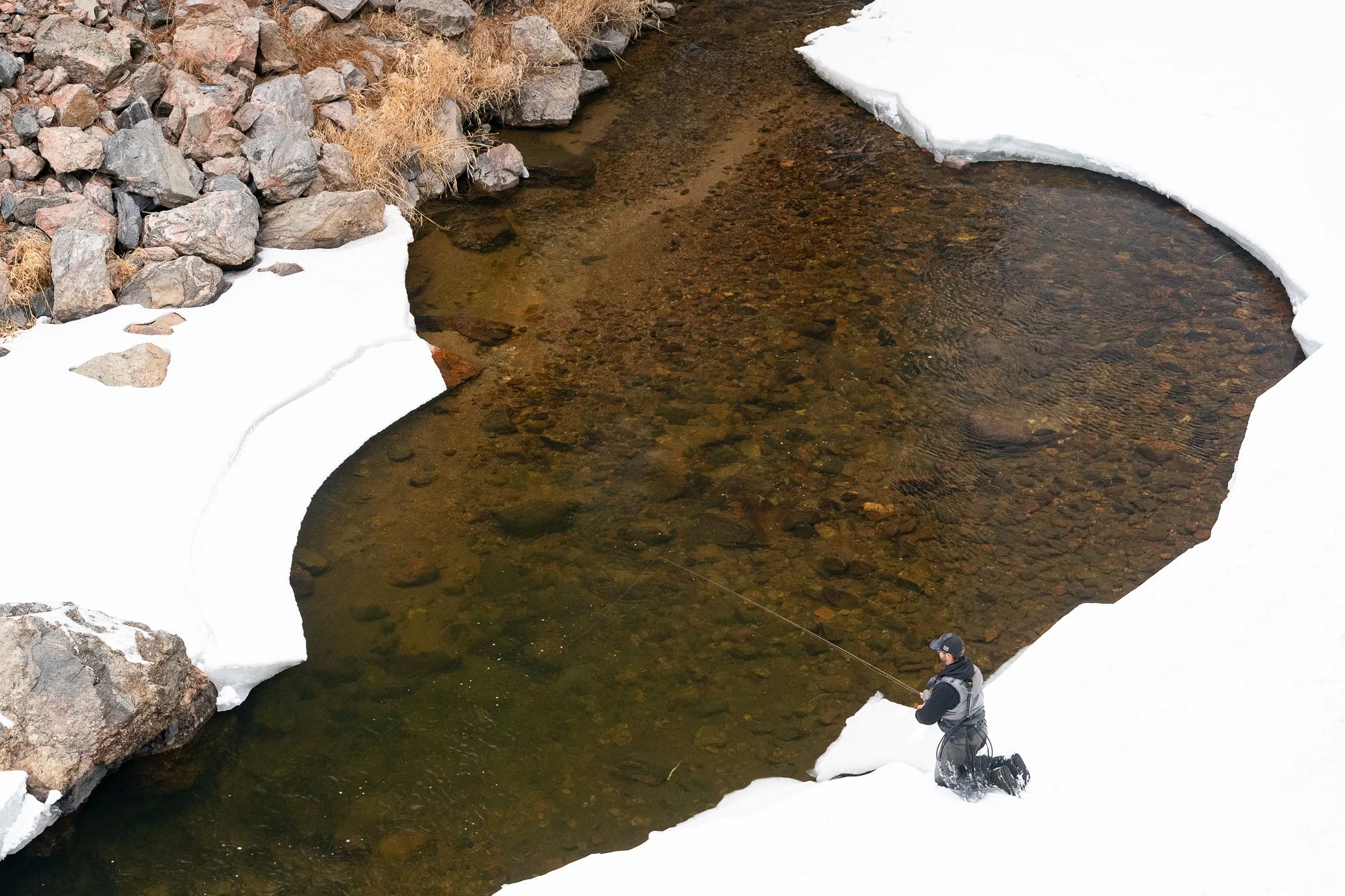 Photograph of a fly fisherman kneeling on ice while casting into the Big Thompson River near Estes Park, Colorado.