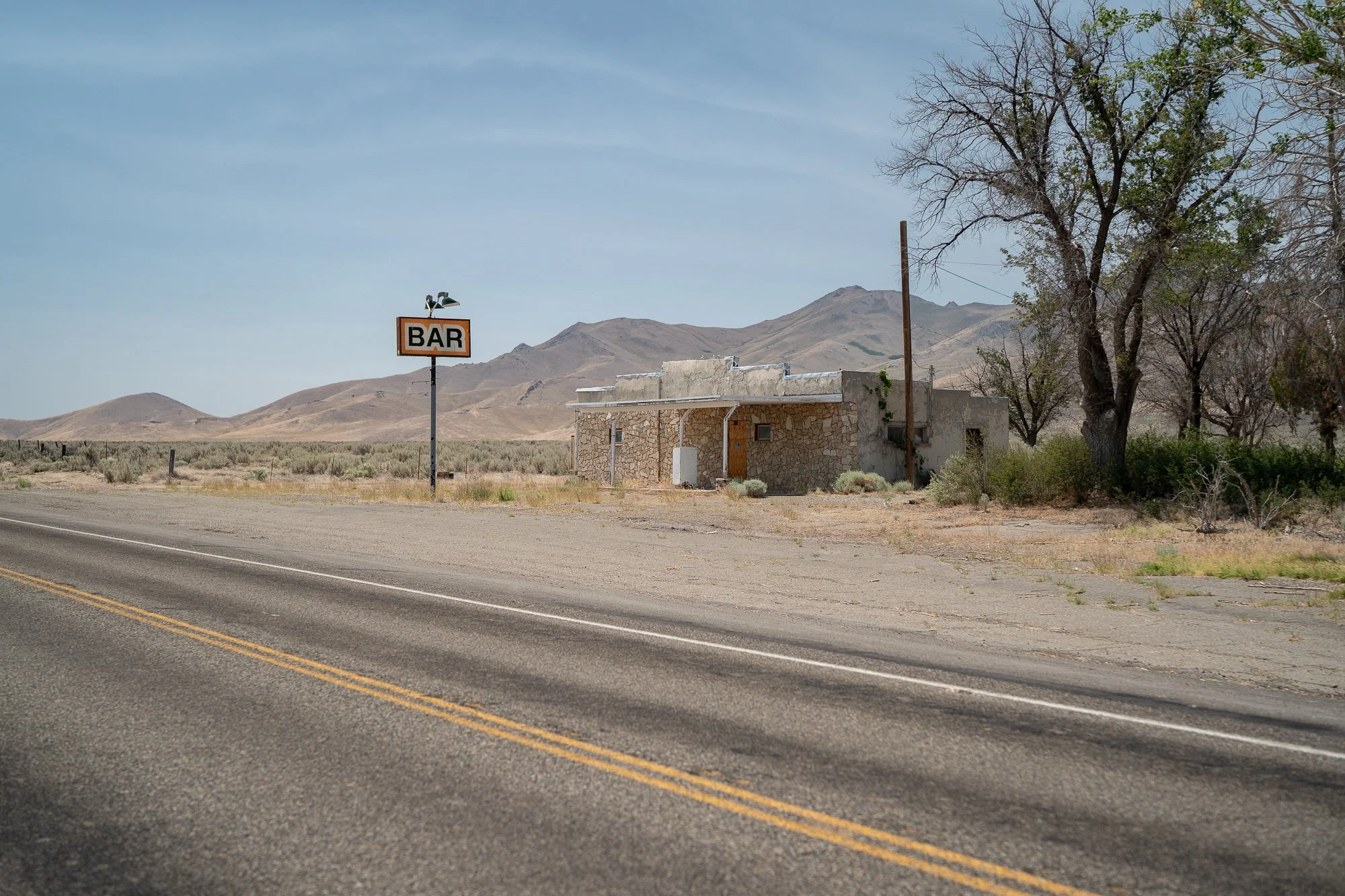 Small stone building with a roadside Bar sign on a post alongside a two-lane Nevada highway, dry trees and desert mountains visible behind it.