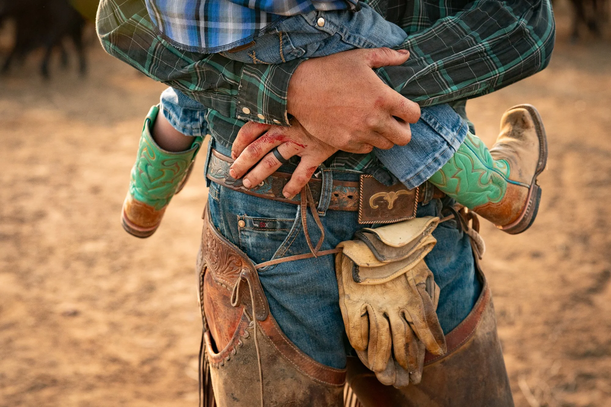 Cowboy holding small child wearing green boots with visible dirt and worn chaps detail