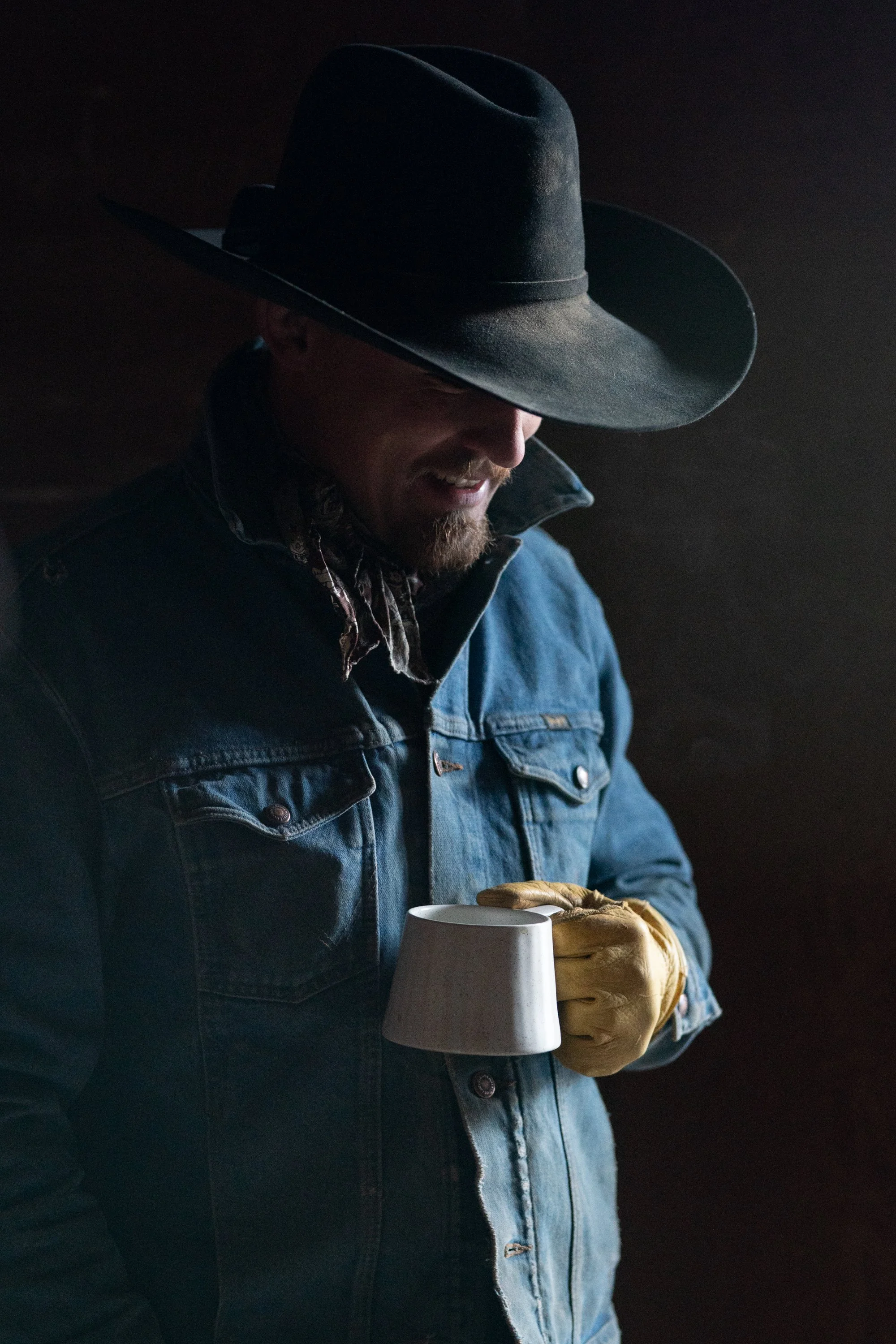 A cowboy wearing a denim jacket and gloves holds a coffee mug inside a barn at Silver Spur Ranch