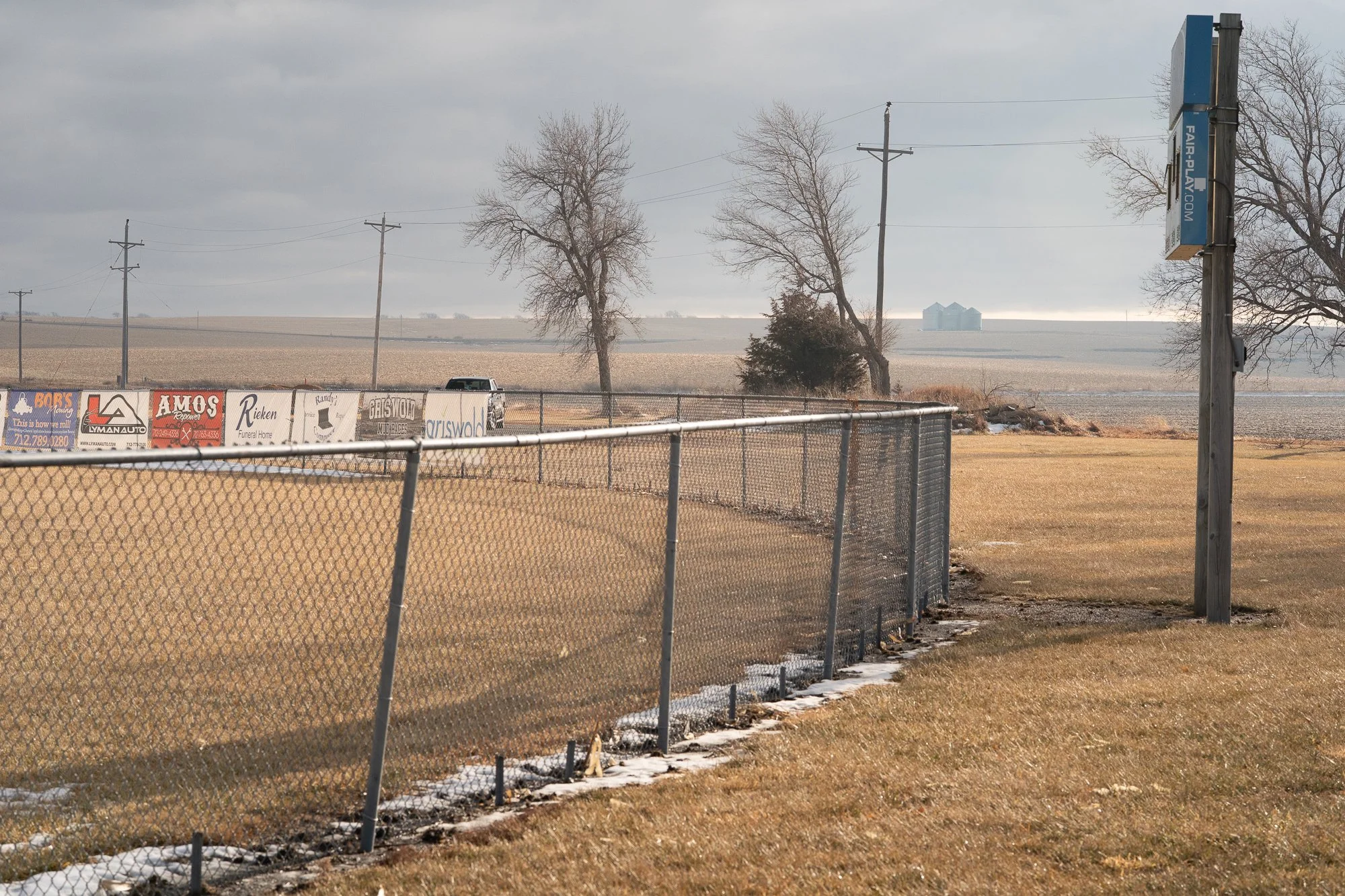 Chain-link fence in front of a ballfield, with farmland, trees, utility poles, and grain silos beyond it.