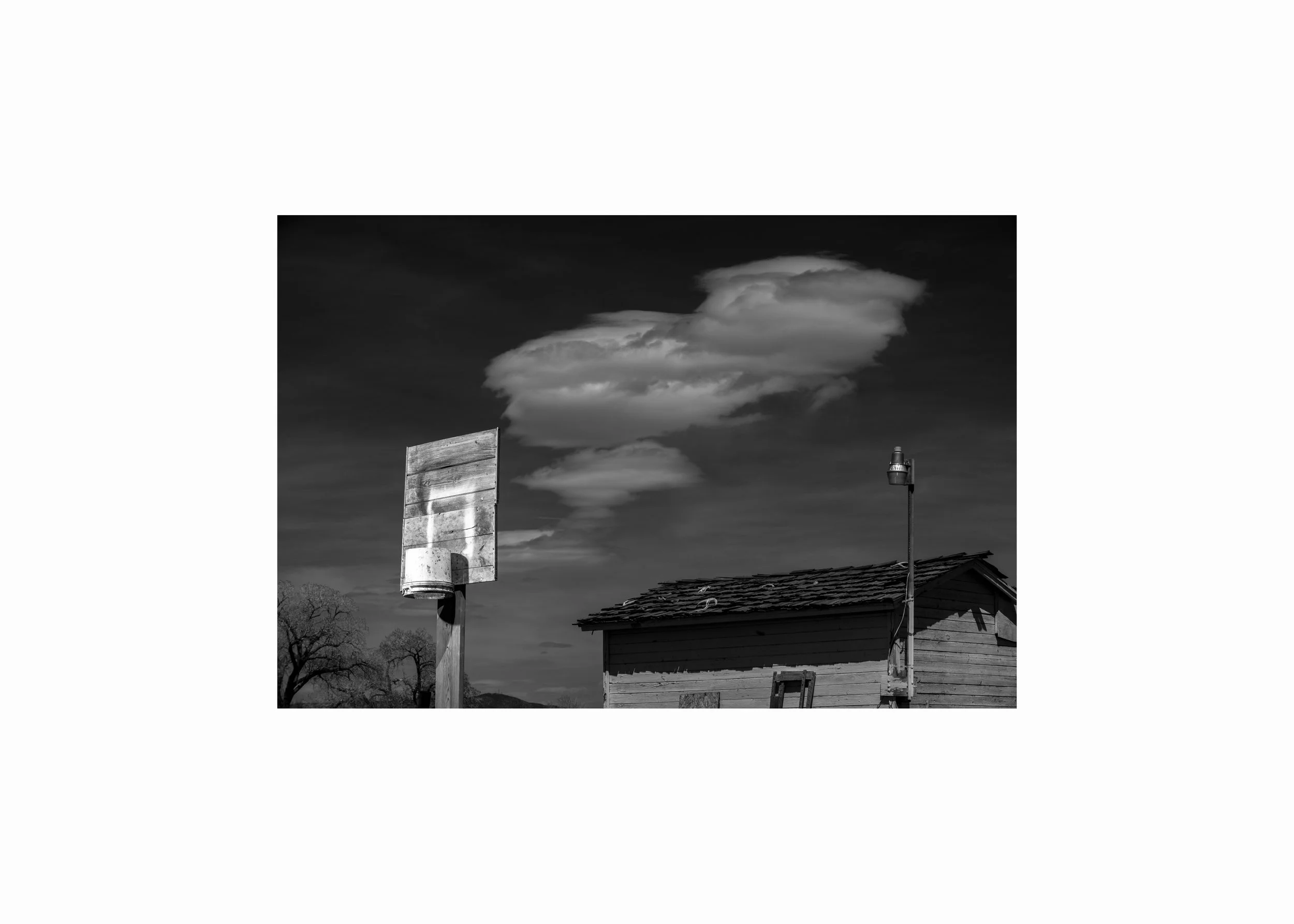 Black and white photograph of a homemade basketball hoop beside a weathered rural building under a dramatic sky.
