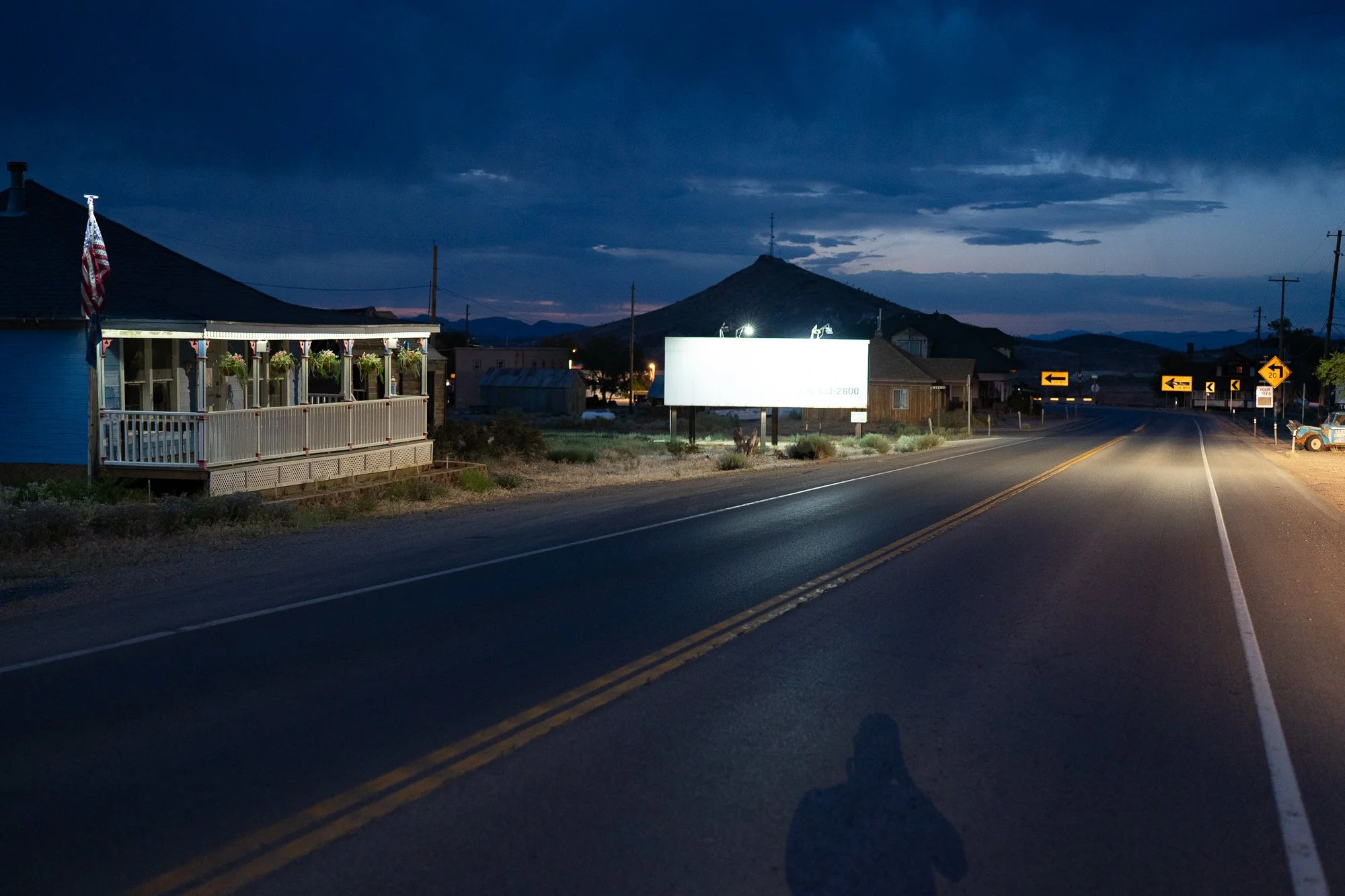 Nevada small town highway at night with a lit blank billboard, a Victorian-style house with a front porch, directional road signs, and a mountain silhouette.