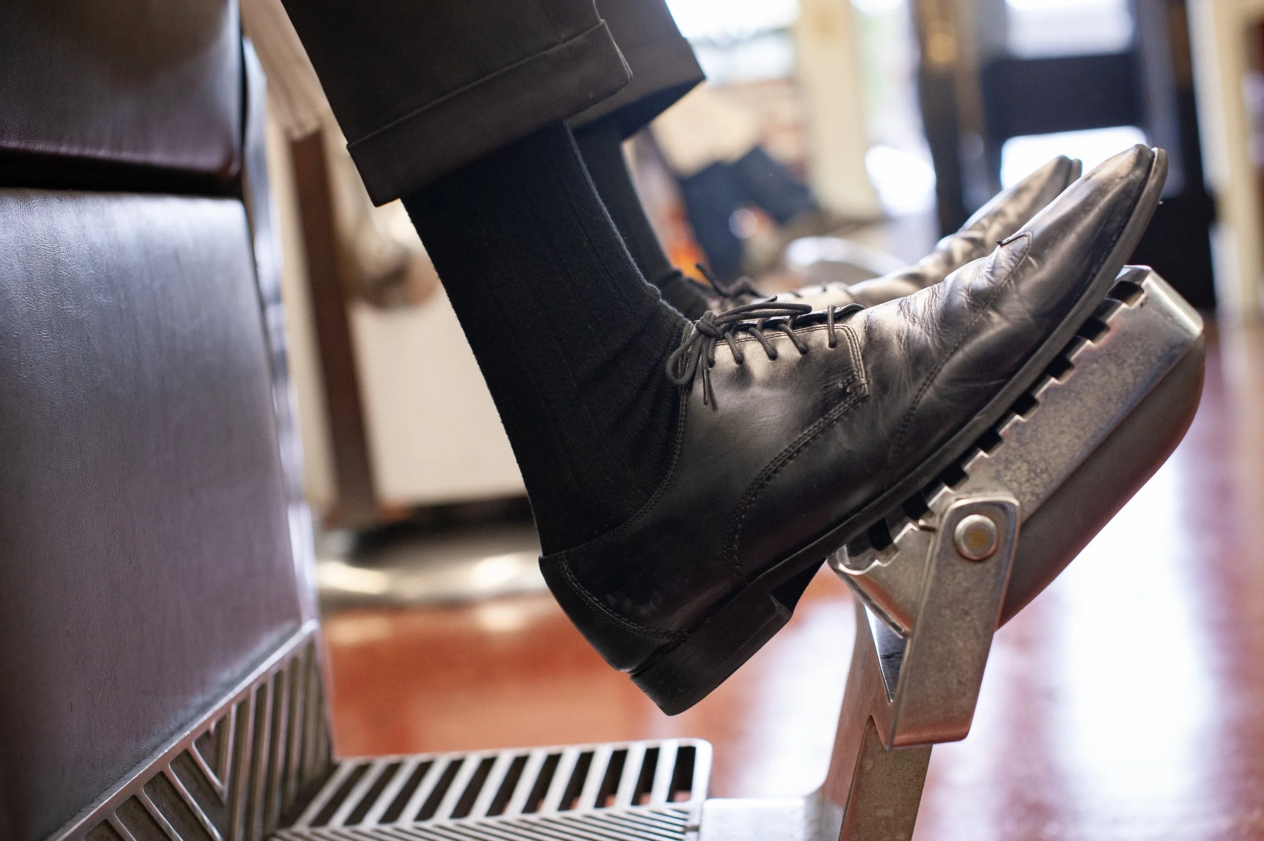Close-up of a polished black leather oxford resting on the chrome footrest of a vintage barber chair at Sweeney Todd's Barbershop
