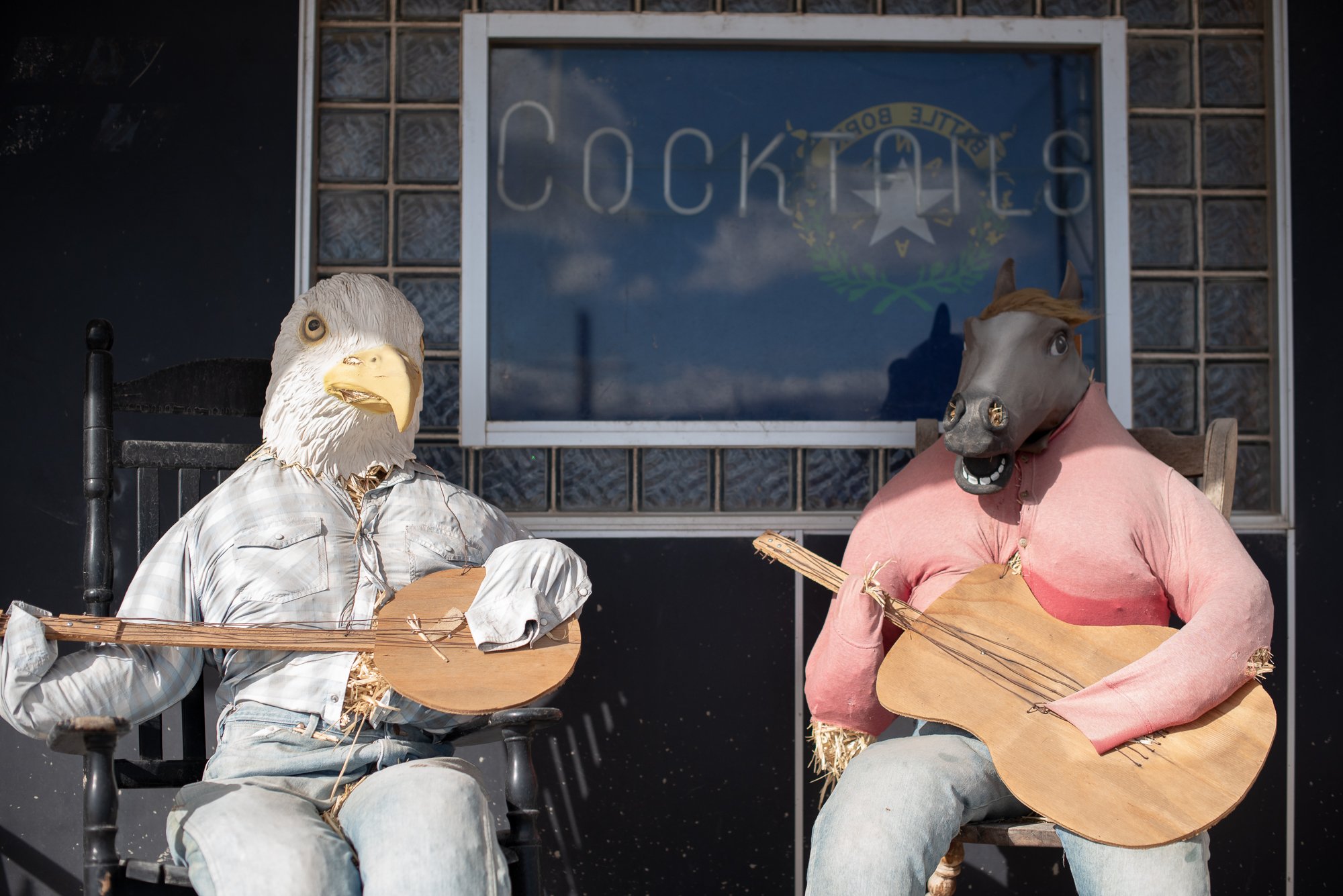 Two scarecrow-style figures with animal masks holding guitars seated outside a Nevada bar with a Cocktails sign visible in the window behind them.