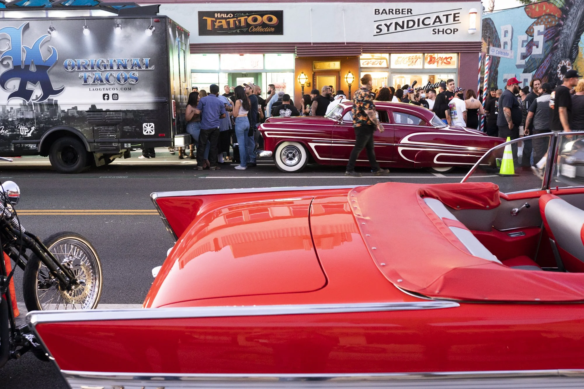 Red and maroon lowriders parked outside Syndicate Barber Shop in Long Beach during a 20th anniversary street celebration with a large crowd and taco truck