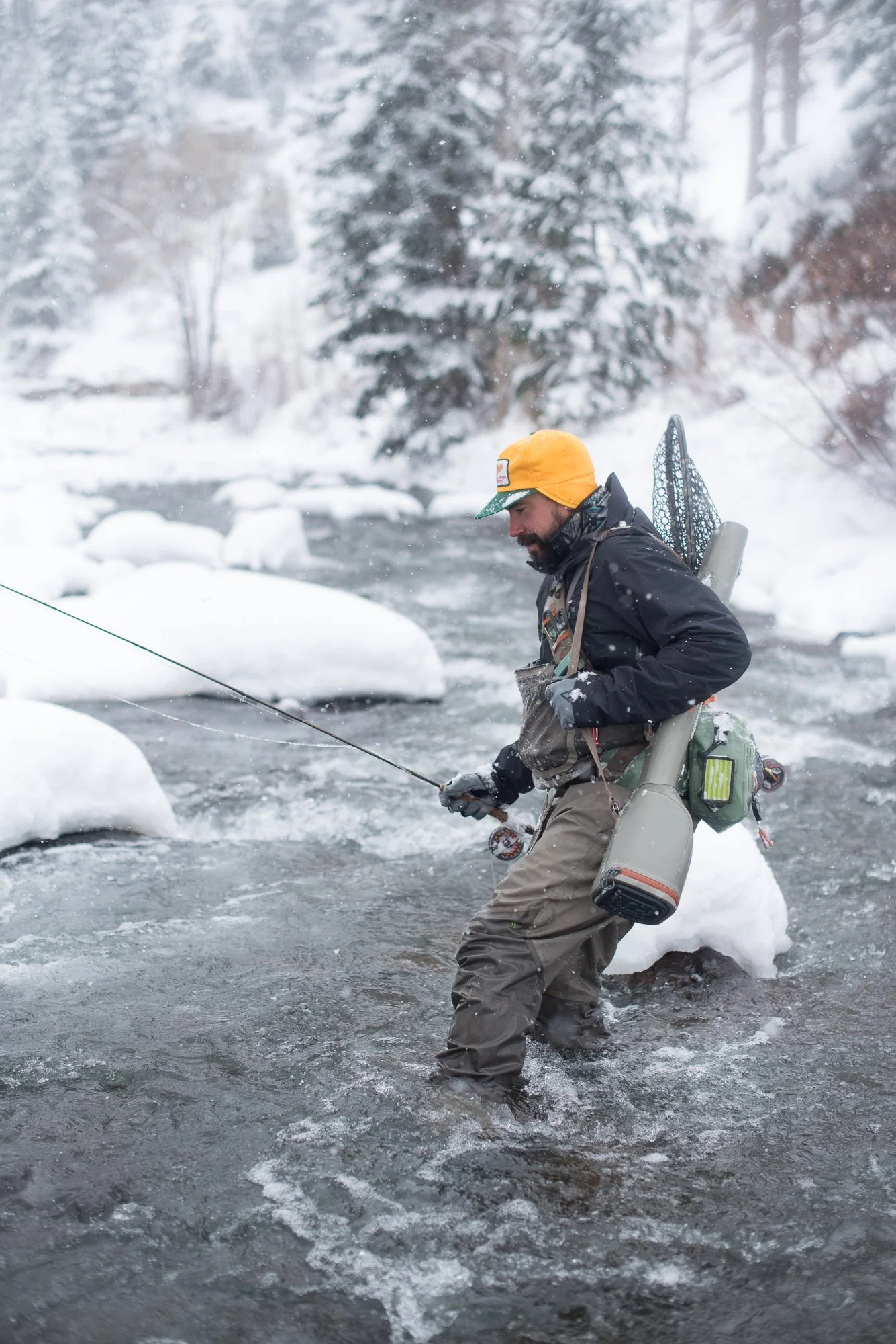 A fisherman wades through Clear Creek in winter with snow falling and gear on his back.