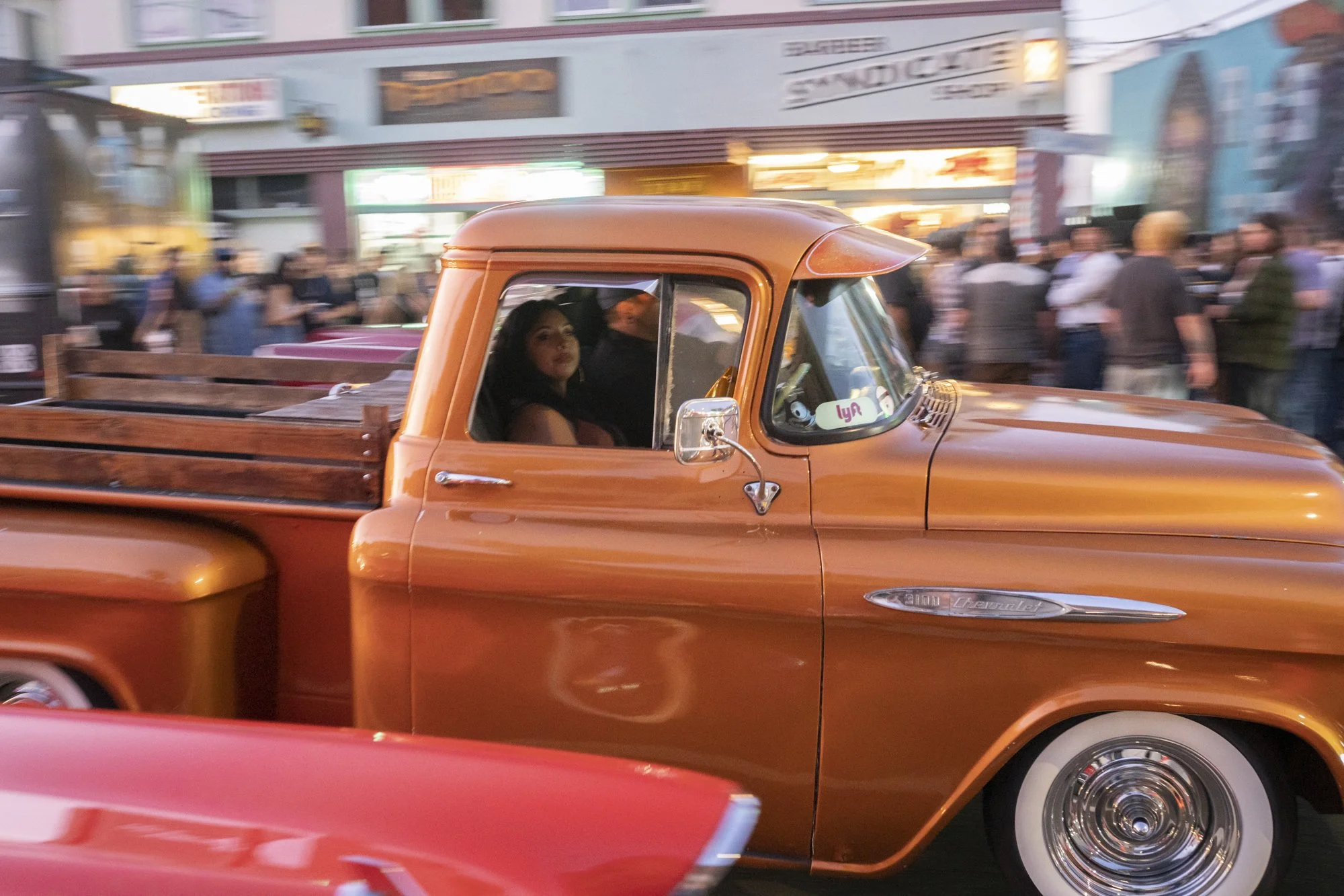 Copper-toned vintage Chevrolet 3100 pickup truck cruising past Syndicate Barber Shop with a woman in the passenger seat and a blurred crowd in the background