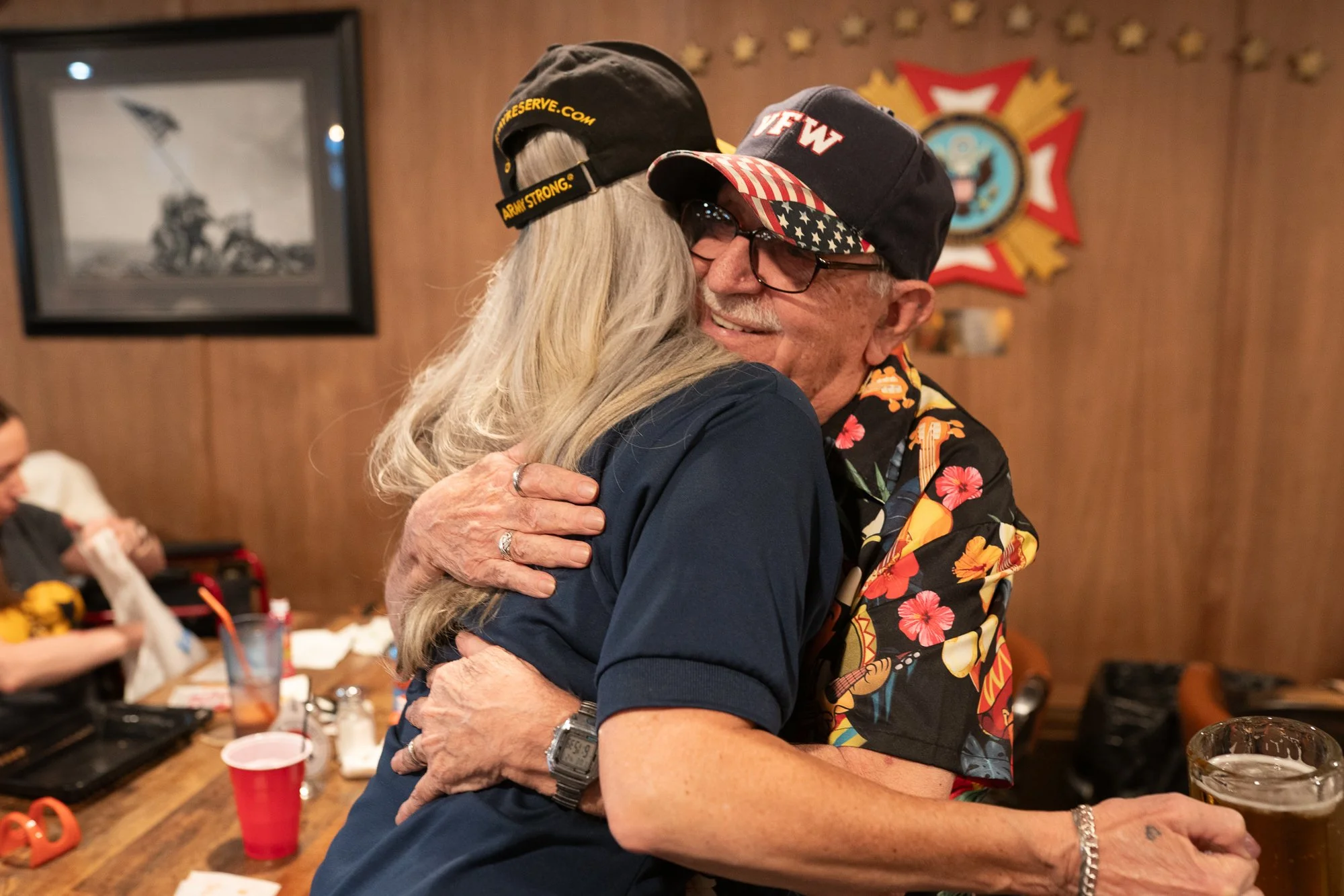 Two people embracing inside a VFW hall with a VFW hat and wall memorabilia visible