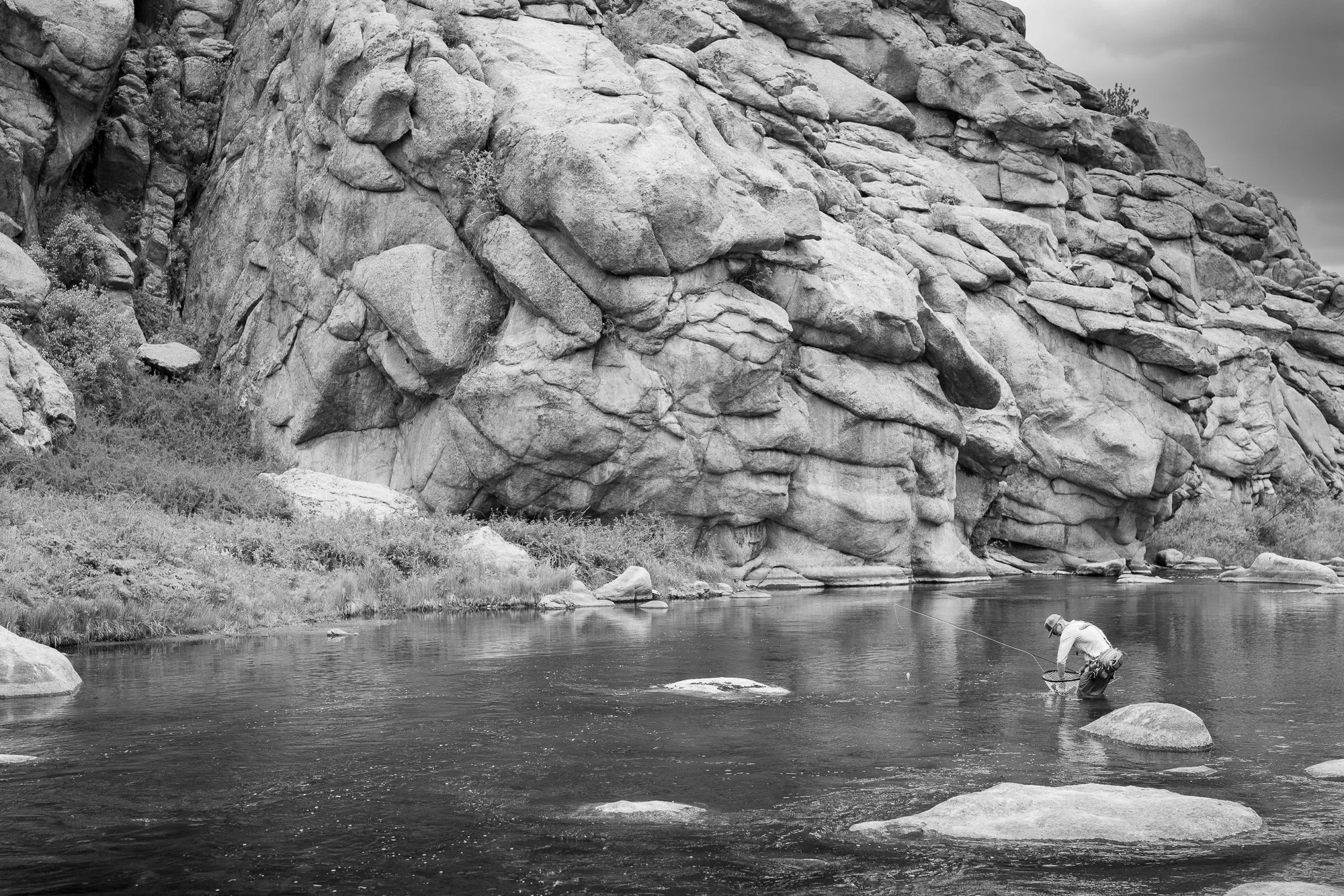 Black and white photograph of a fly fisherman netting a trout in front of large granite walls