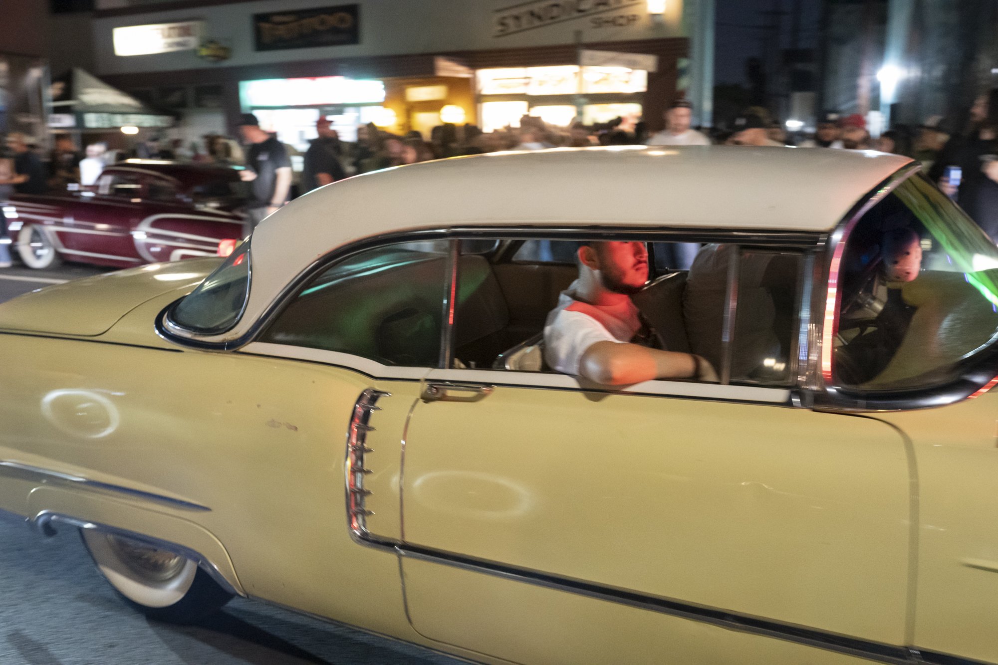 Cream-colored lowrider sedan with white vinyl roof cruising past Syndicate Barber Shop at night with a maroon lowrider visible behind it
