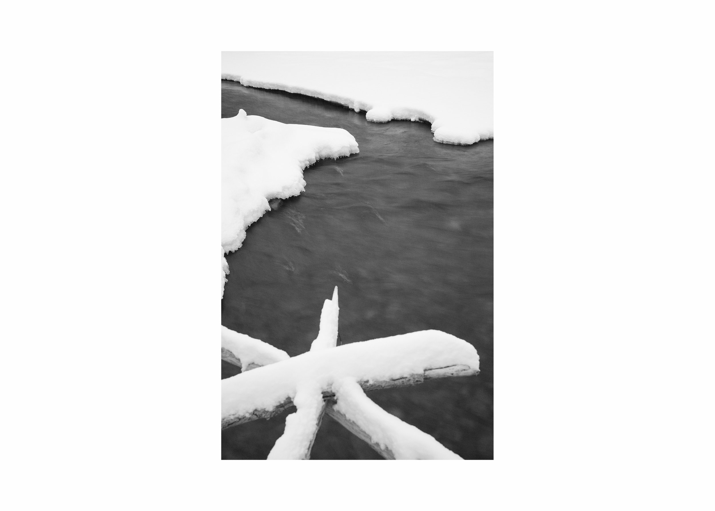 Minimalist black and white photograph of a partially frozen river, with smooth flowing water framed by snow-covered ice and branches, evoking winter stillness and natural calm.