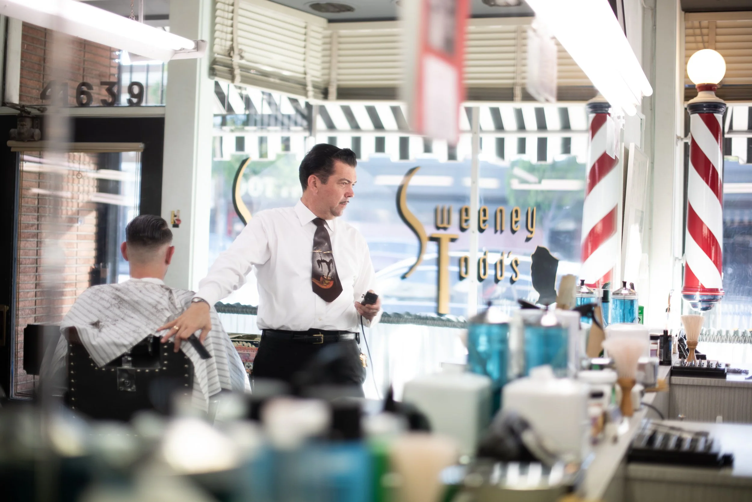 Barber in white shirt and tie pausing with clippers to assess a client's cut at Sweeney Todd's Barbershop, framed by gold storefront lettering and barber pole in the window