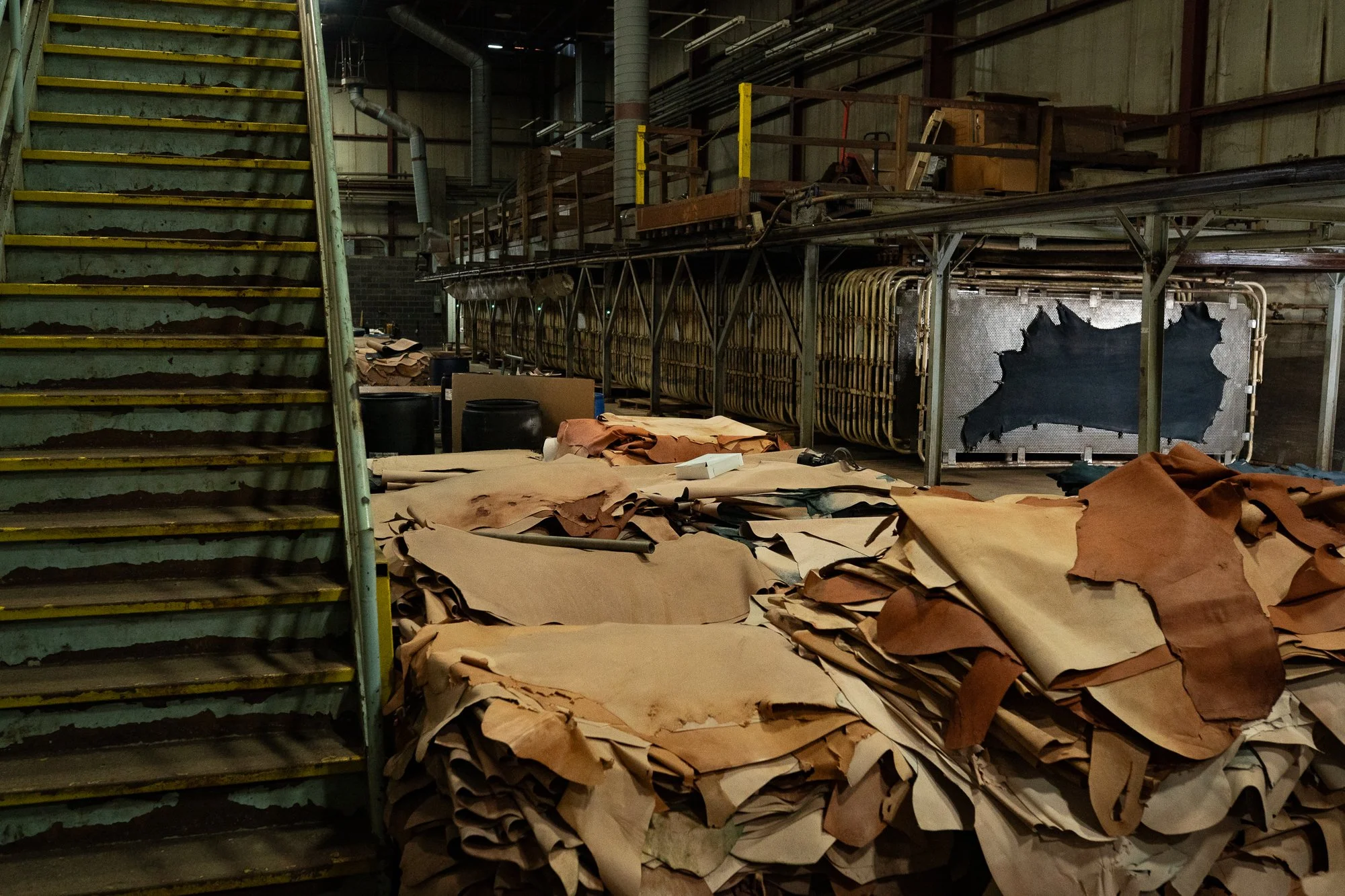 Stacks of brown leather hides fill a production area alongside machinery, carts, and work surfaces