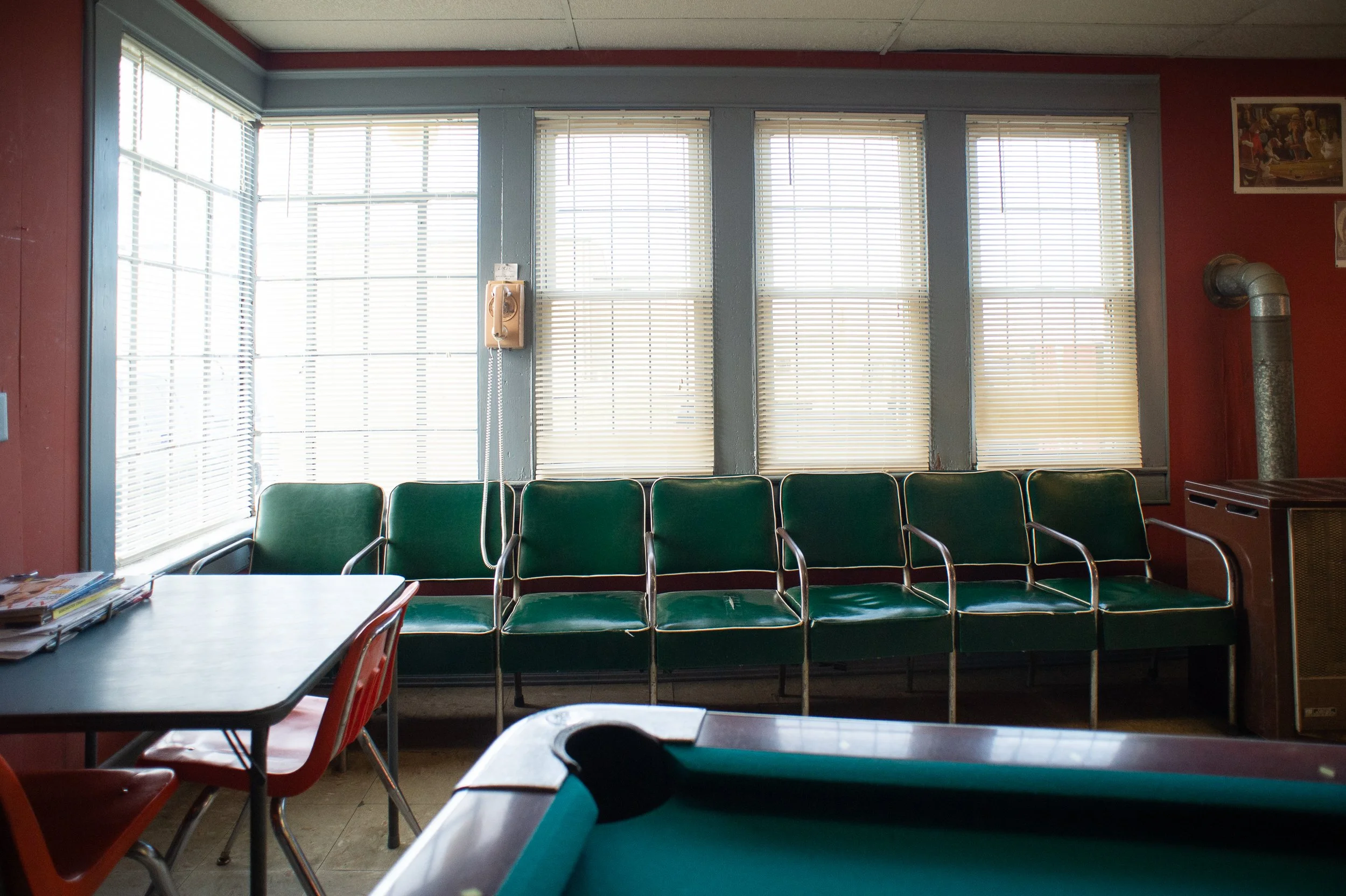 Row of green waiting chairs beneath large windows with blinds inside a barbershop in Maryland