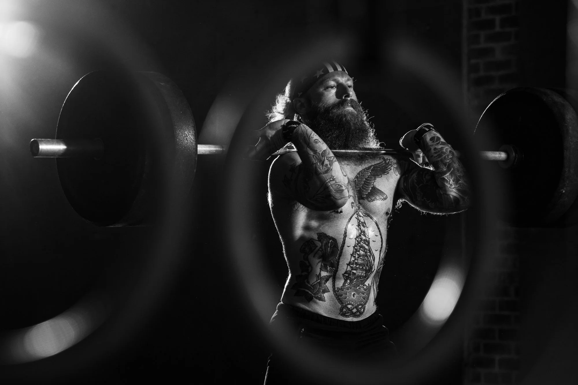 Bearded CrossFit athlete holding a loaded barbell in the front rack position inside a gritty gym.
