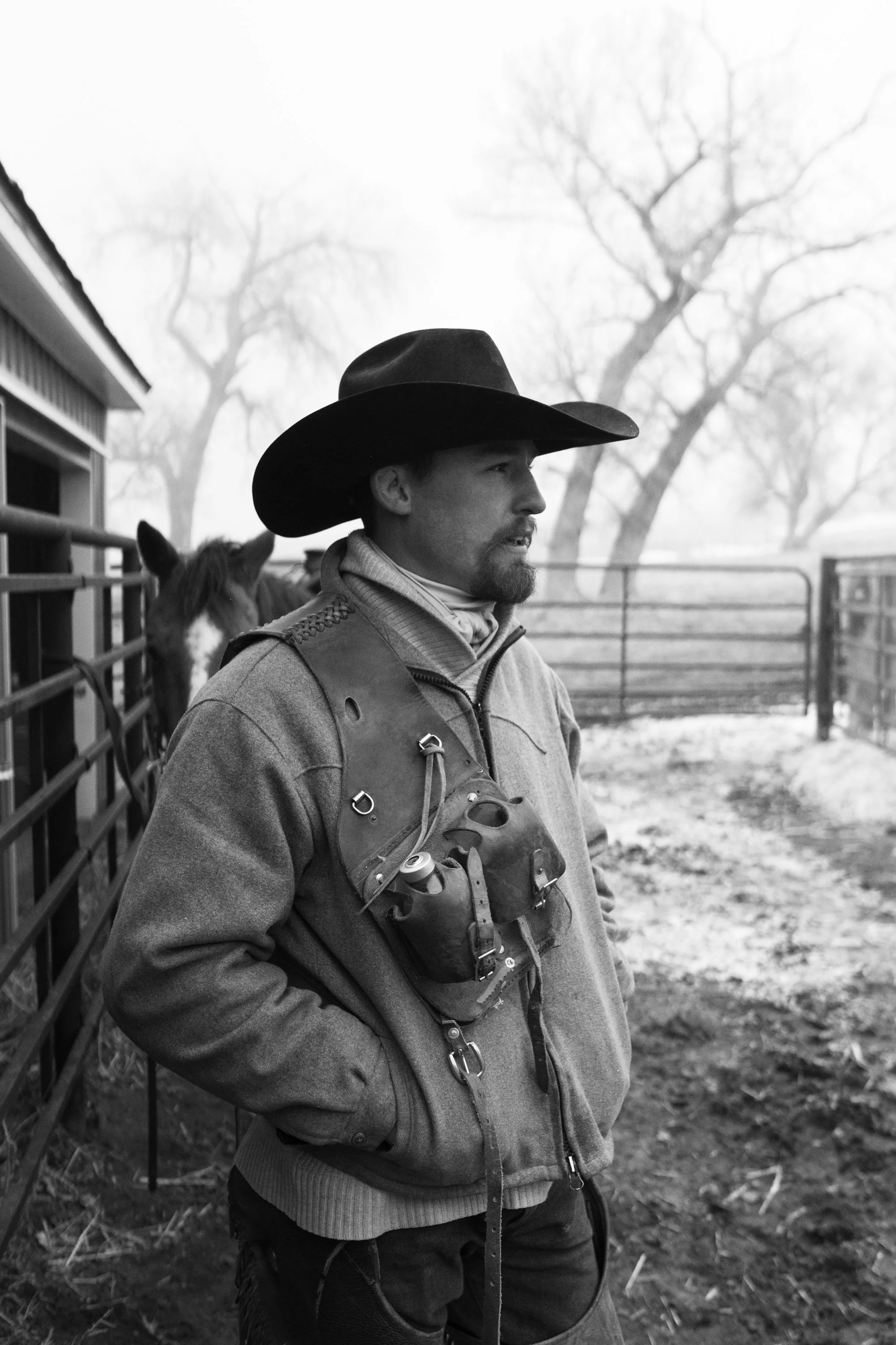 A cowboy stands beside the barn during calving season at Silver Spur Ranch with a horse waiting behind him