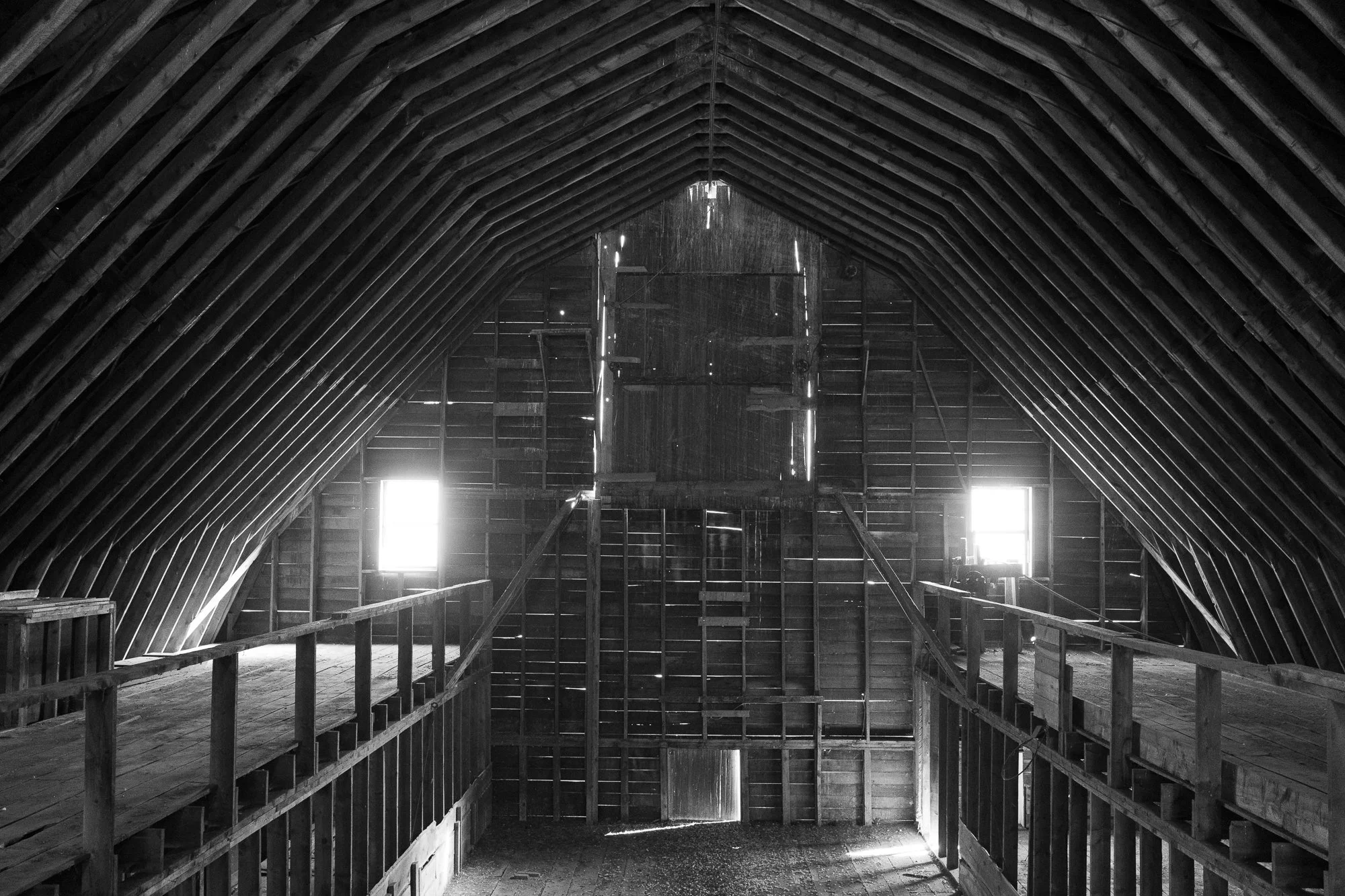Black-and-white view inside a historic barn showing exposed timber rafters, wooden loft walkways, plank walls and daylight entering through windows and gaps in the siding.
