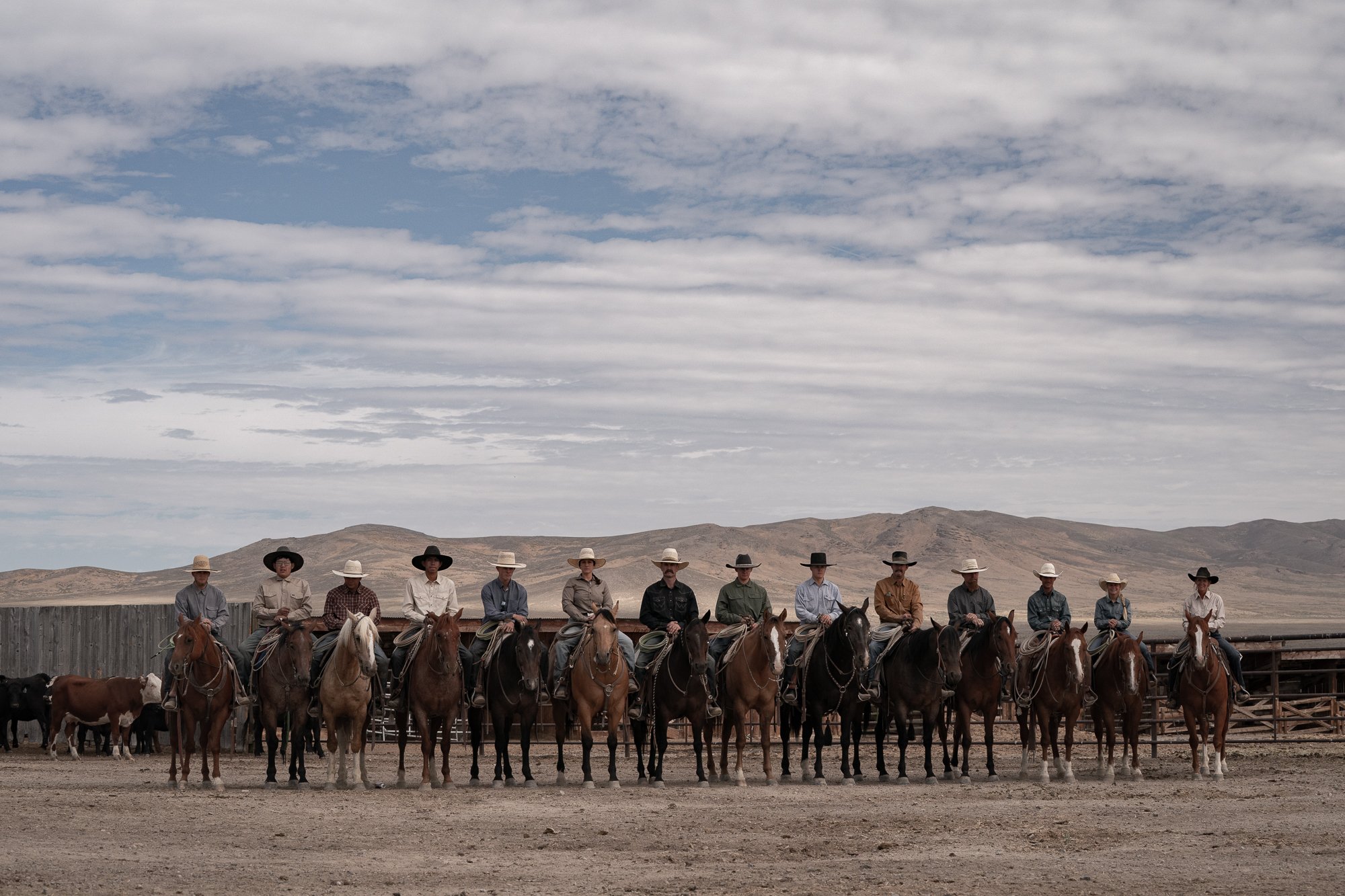 Line of buckaroos on horseback in front of a ranch corral, with cattle and dry mountains in the background.