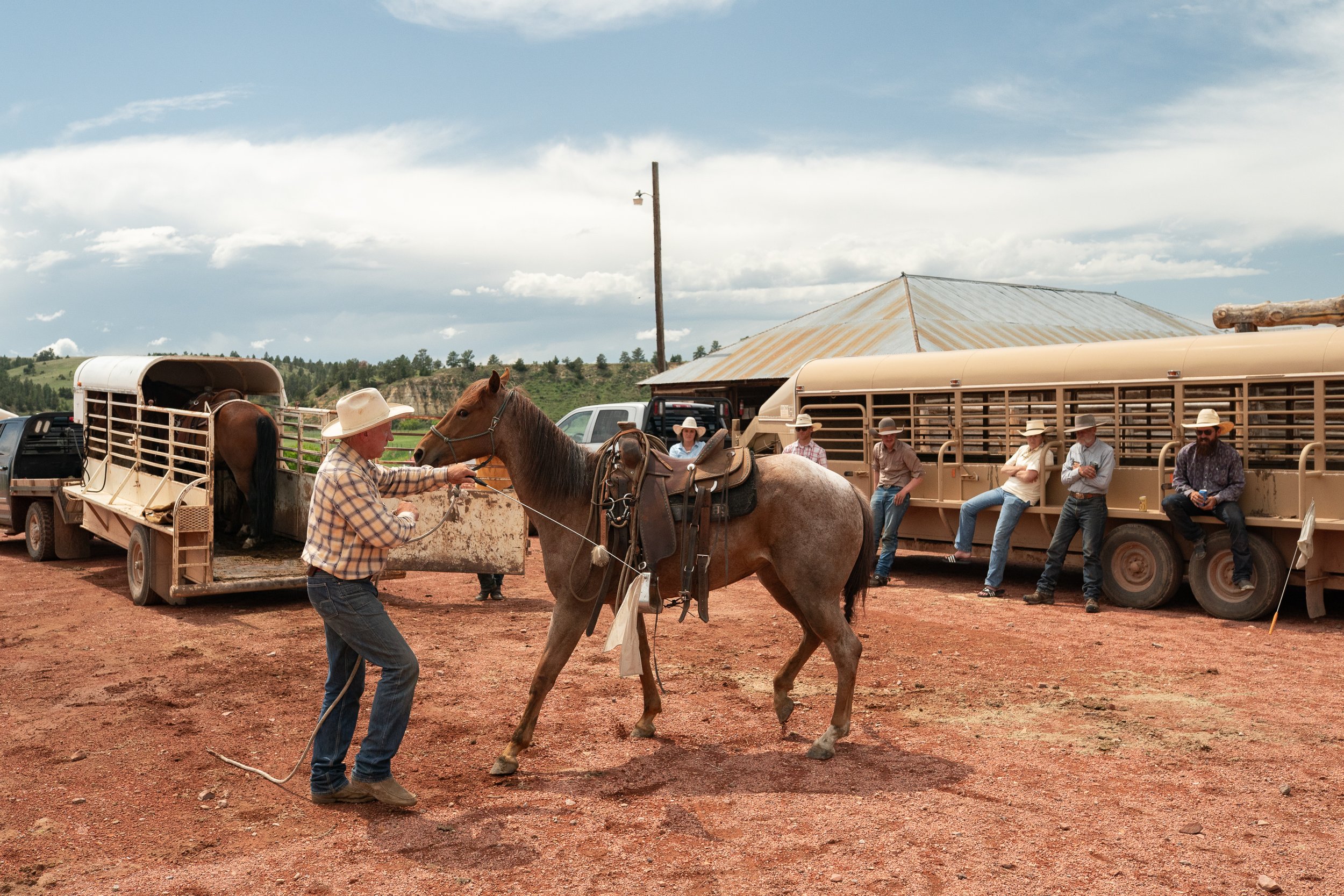 Buck Brannaman working a horse with a lead rope while cowboys sit and watch near trailers on a Montana ranch