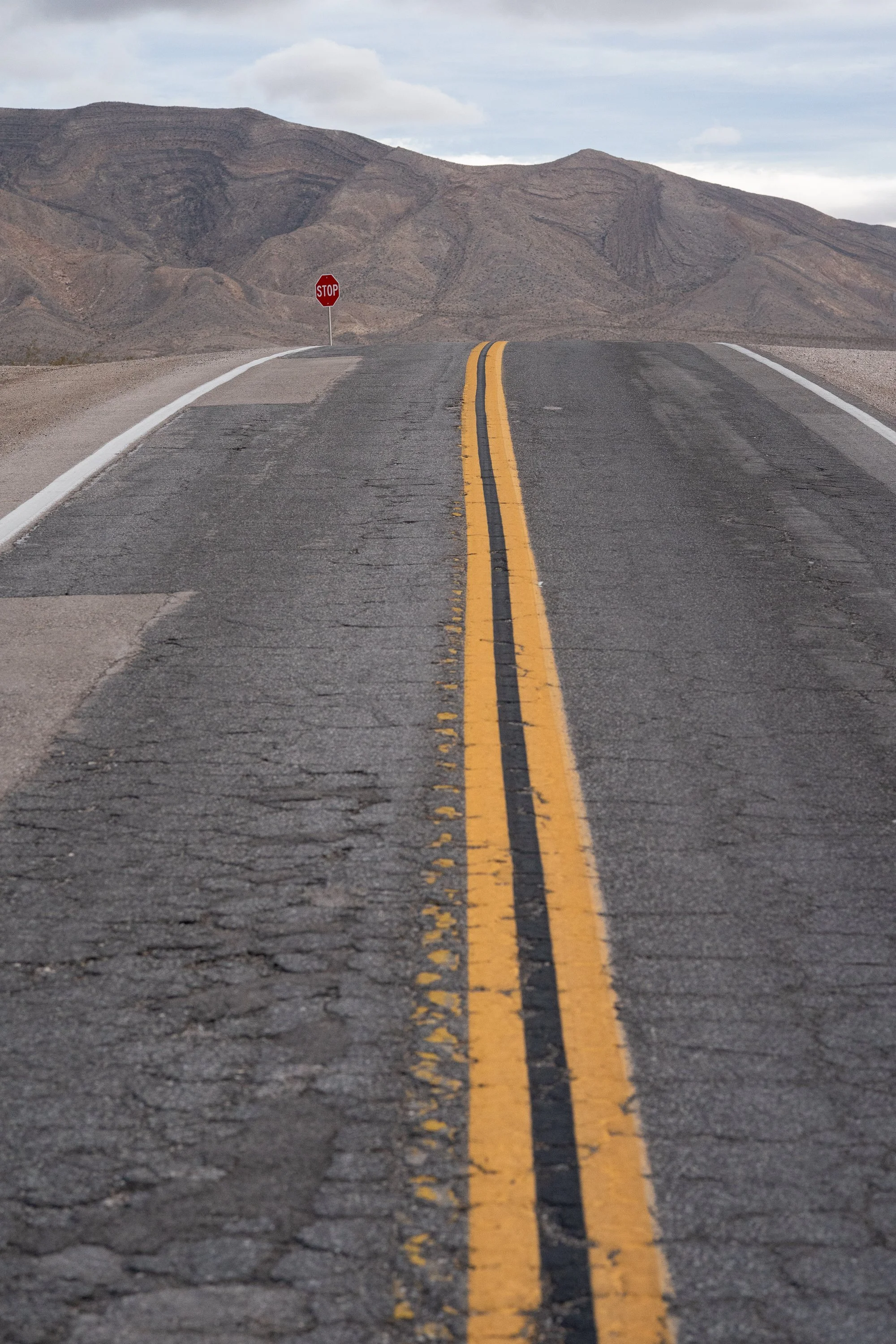 Low-angle view of a cracked and patched Nevada desert highway with double yellow center lines and a lone stop sign visible in the middle distance against a dry mountain.