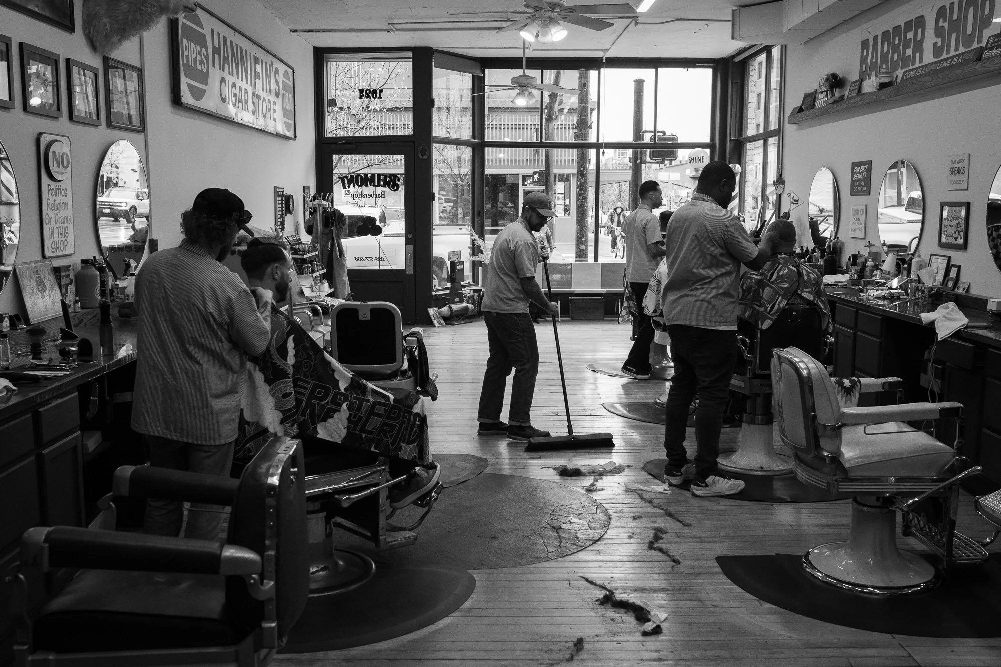 wide black and white interior of Belmont Barbershop in Boise Idaho with barbers cutting hair and sweeping the floor