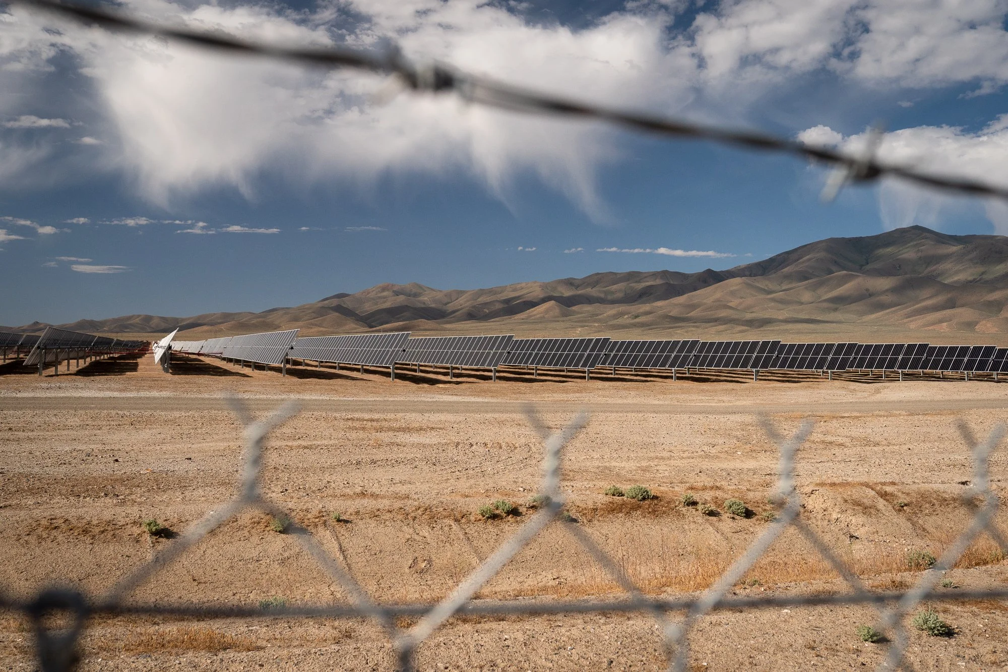 Rows of solar panels behind a chain-link fence in the Nevada desert, with dry scrubland and brown mountain ranges visible in the background.