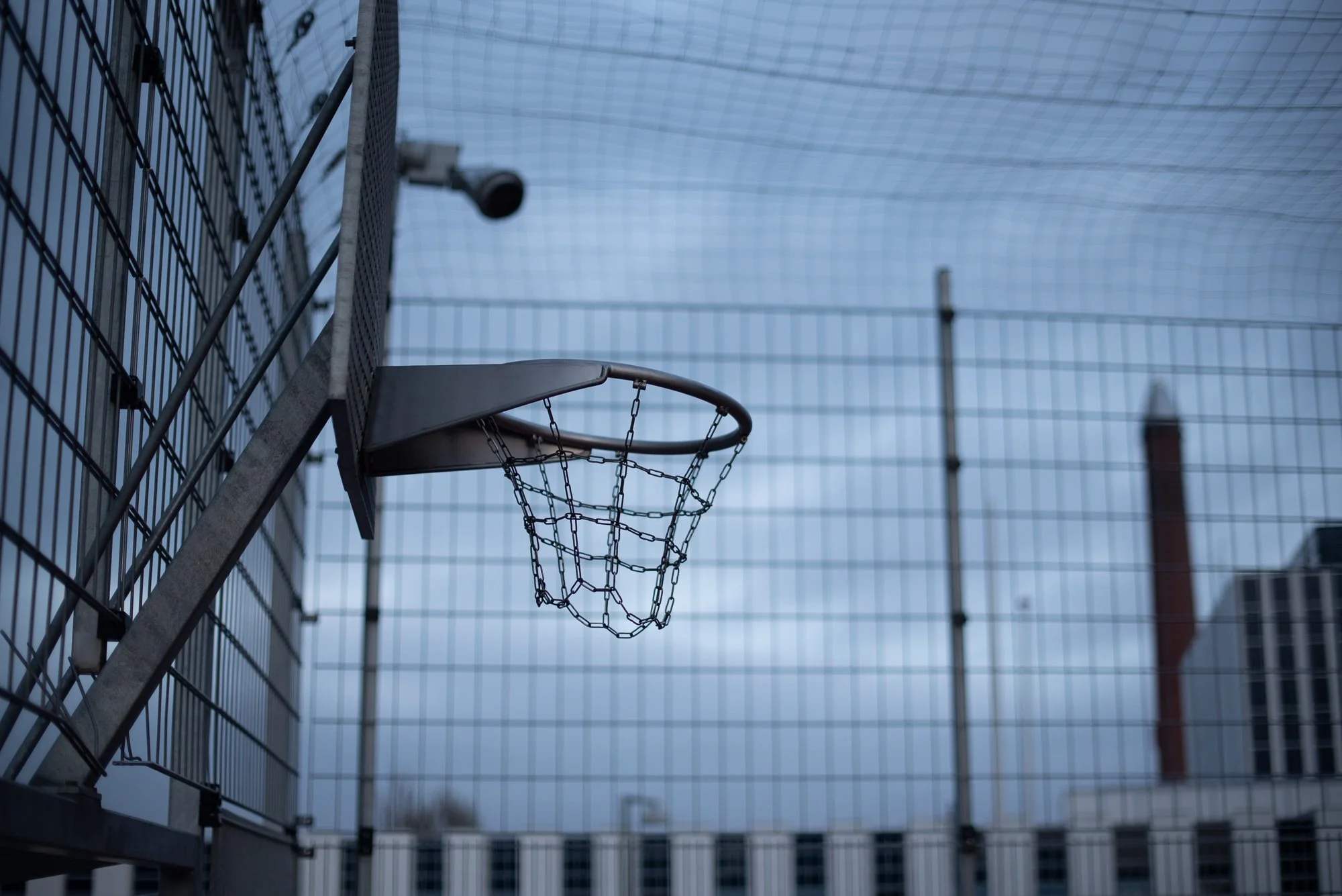 Outdoor basketball hoop photographed at dusk behind fencing on a European public court.
