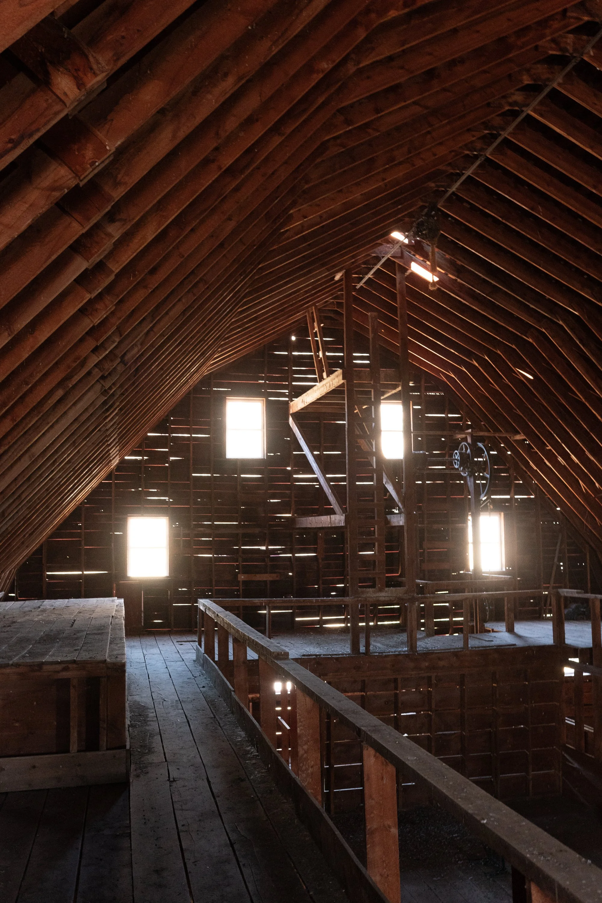 Interior of an old wooden ranch barn with exposed roof beams, elevated walkways, a central pulley mechanism and sunlight coming through gaps in the plank walls.