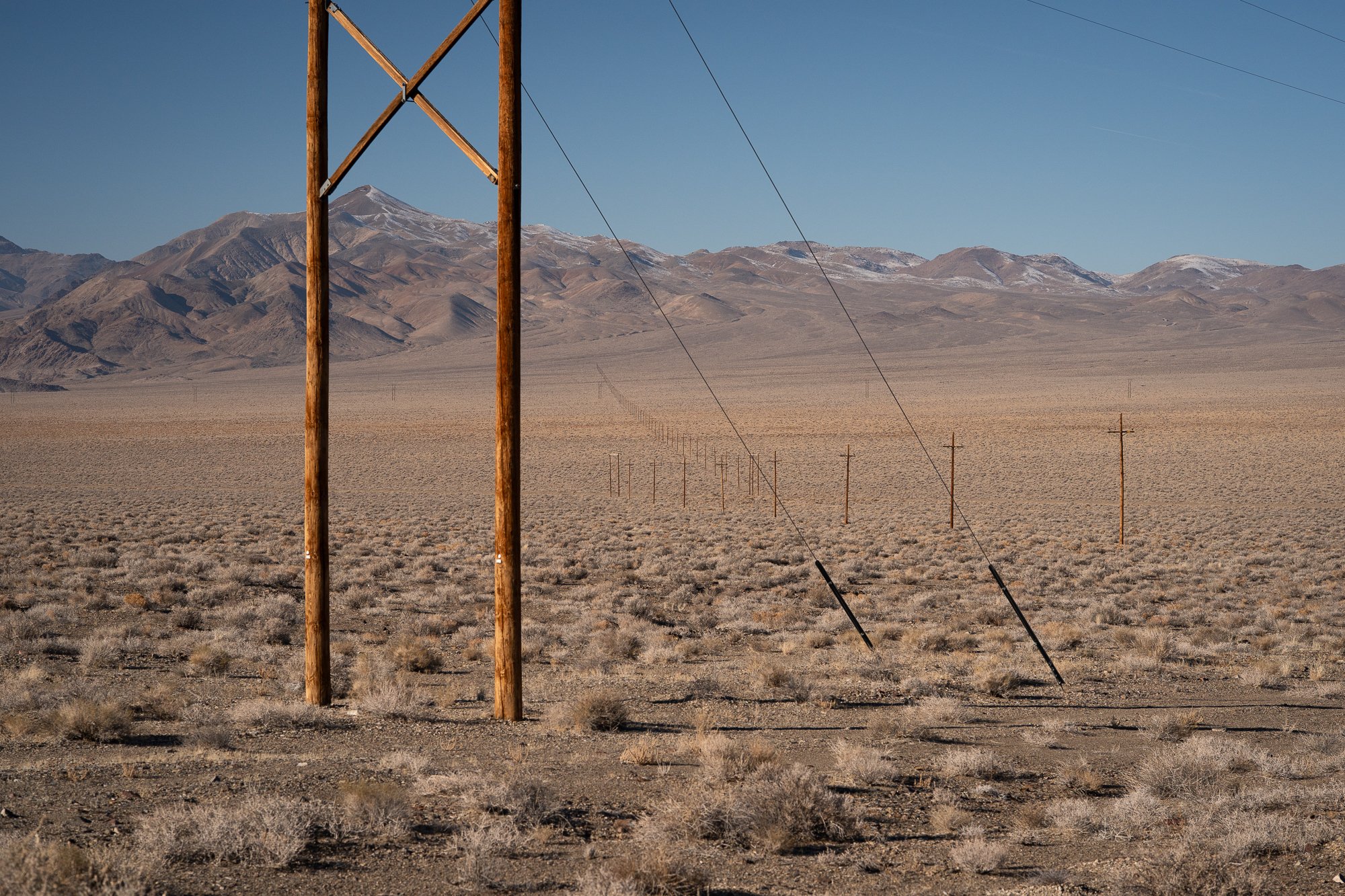 Double wooden utility poles with cross-arms in the Nevada desert foreground, with a long line of poles receding into sagebrush flats and snow-dusted mountains under a clear blue sky.
