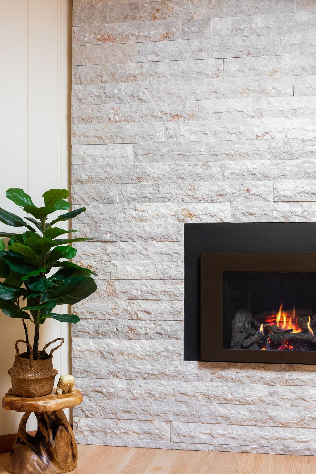Living room corner with a potted plant on a wooden stand, a small decorative elephant, and a fireplace with a black frame, all against a white stone brick wall.