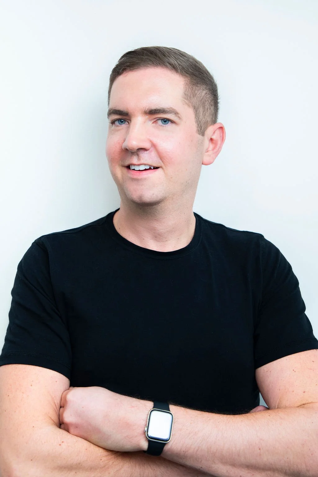 A young man with short brown hair and blue eyes, smiling slightly, wearing a black shirt and a smartwatch on his left wrist, standing against a plain white wall.