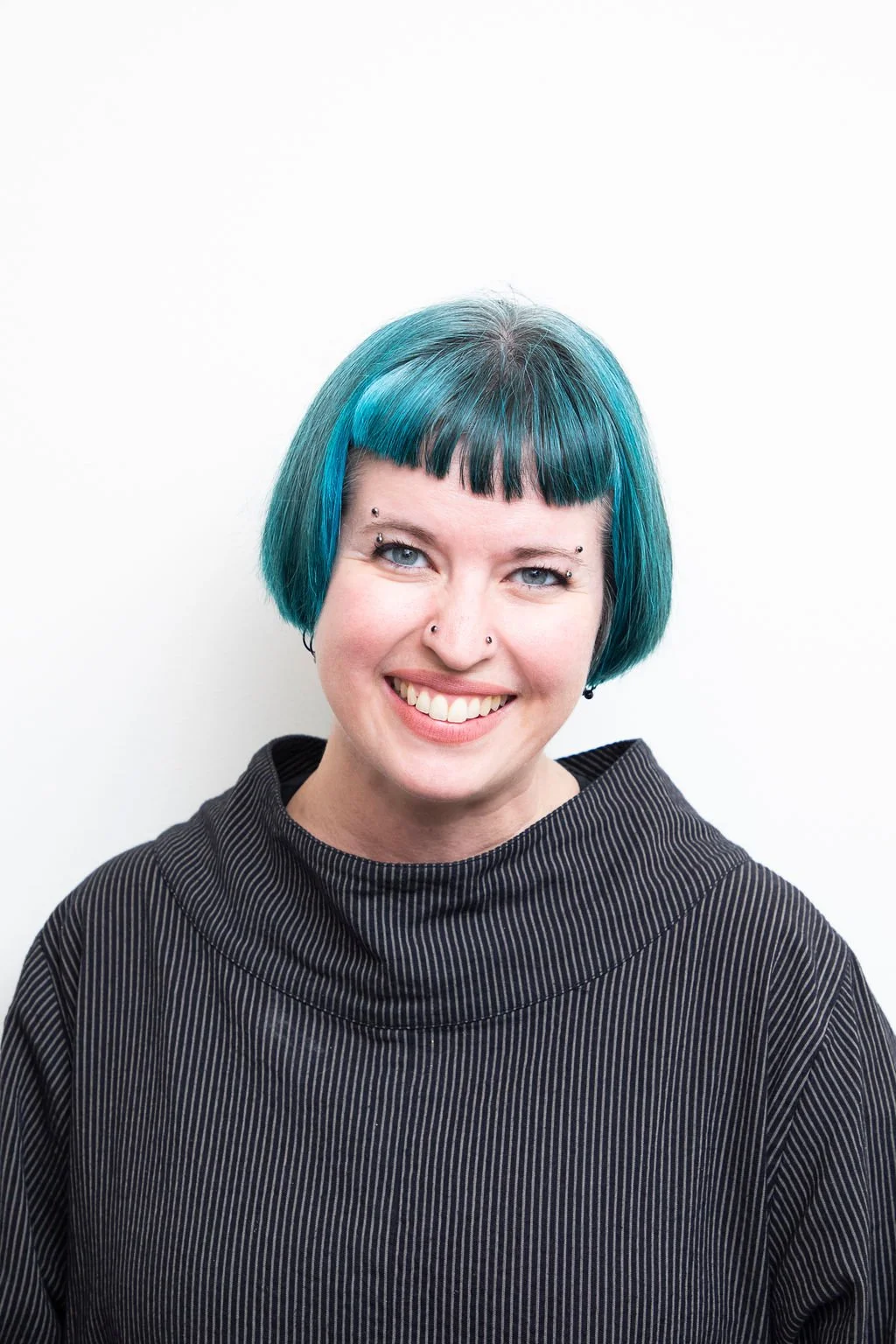A young woman with short, vibrant blue hair, multiple facial piercings, and a wide smile, wearing a black striped top against a plain white background.