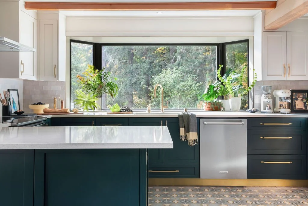 Modern kitchen with white upper cabinets, dark green lower cabinets, a large window above the sink, and various plants on the countertop.
