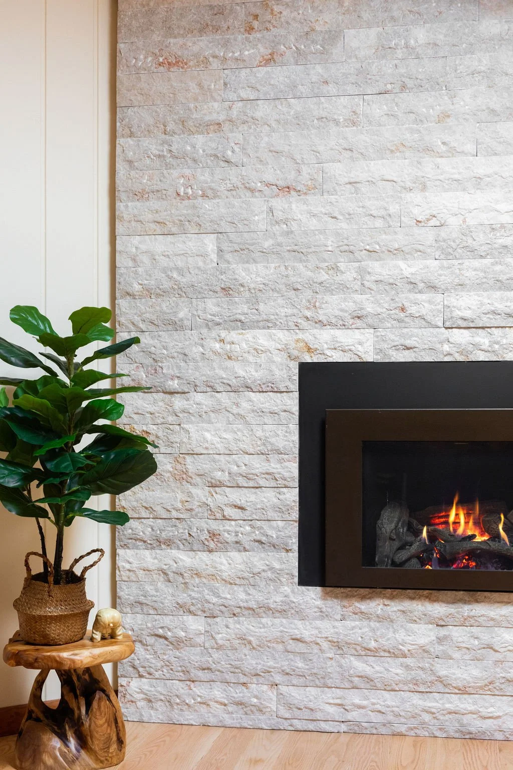 A living room corner with a potted green plant on a natural wood side table, next to a modern fireplace built into a white textured stone wall, with visible flames and fire logs inside.