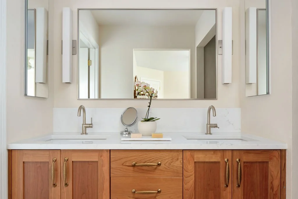 Double bathroom vanity with a marble countertop, two undermount sinks, and modern faucets. Wide mirror above with lighting fixtures on both sides. Centered plant and mirror on tray with soap and accessories.