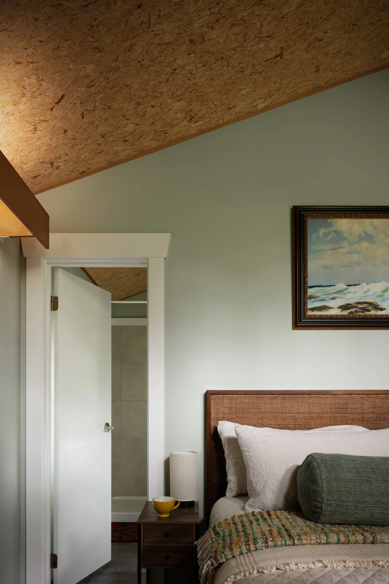 Interior view of a bedroom with a bed, framed landscape painting, nightstand with a lamp and yellow mug, open door revealing a bathroom with shower, and a textured ceiling.