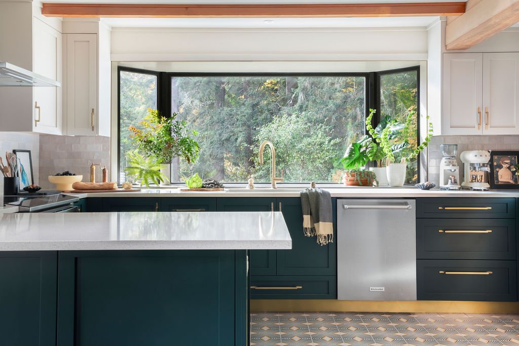 Modern kitchen with large window showing greenery outside, white upper cabinets, dark lower cabinets with gold handles, marble countertop, and various plants and kitchen appliances.