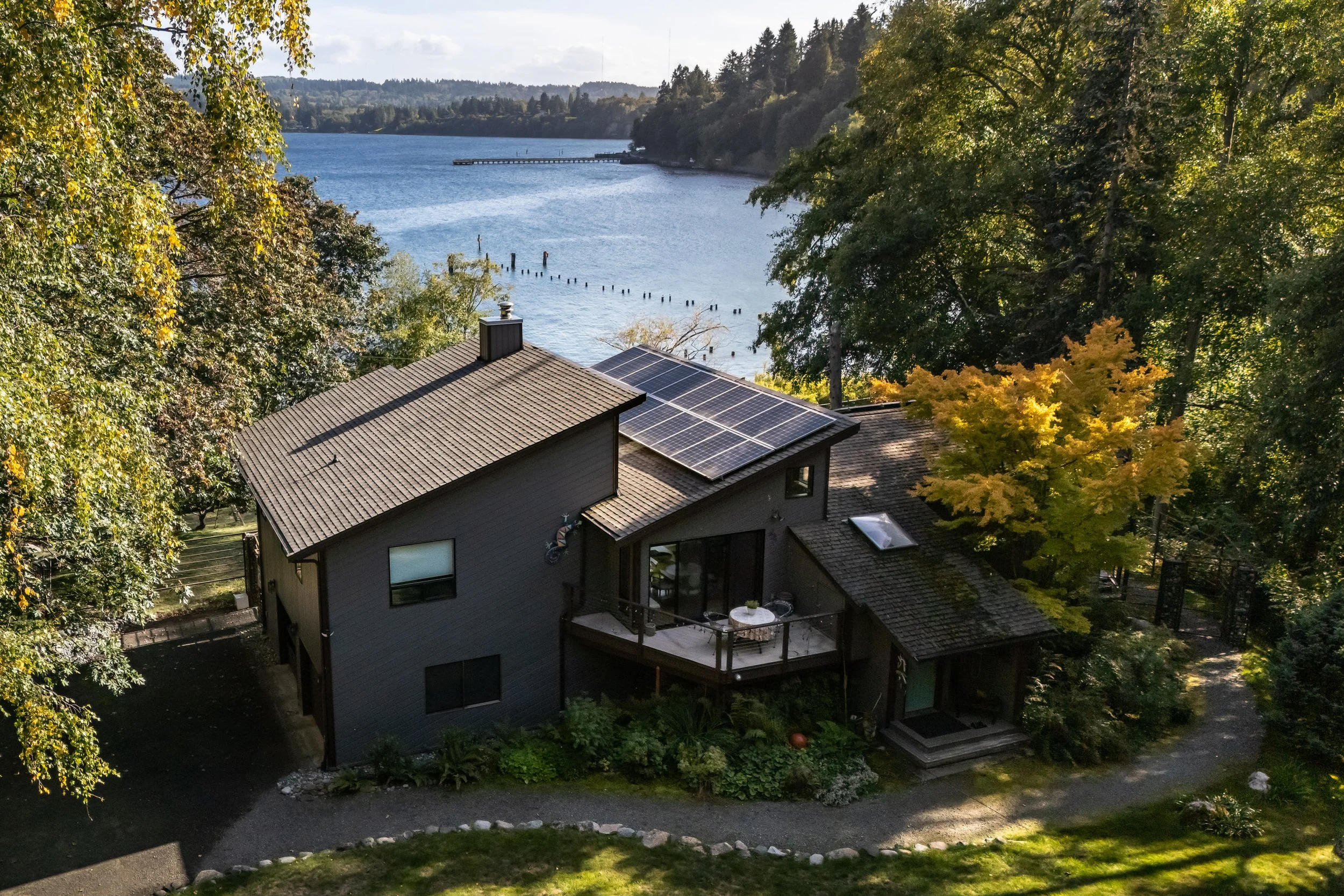 A house by the water with solar panels on the roof, surrounded by trees with green and yellow leaves, and a pathway leading to the entrance.