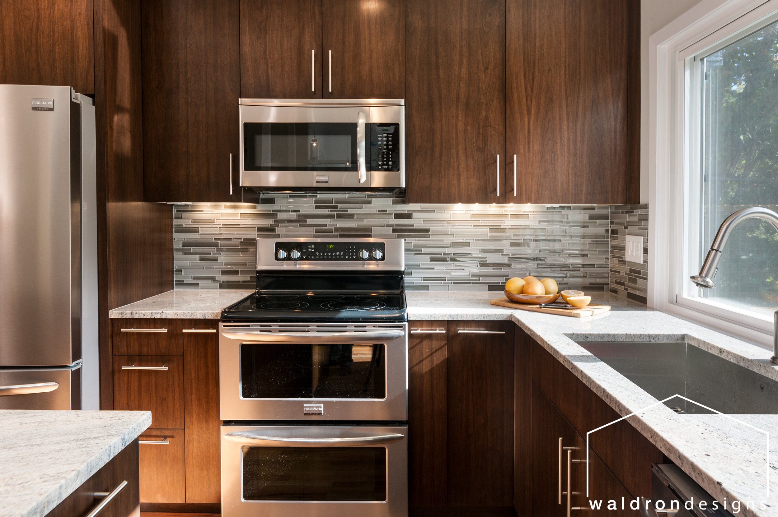 Modern kitchen with wooden cabinets, stainless steel microwave and oven, granite countertops, a gray tile backsplash, and a window with view of trees.