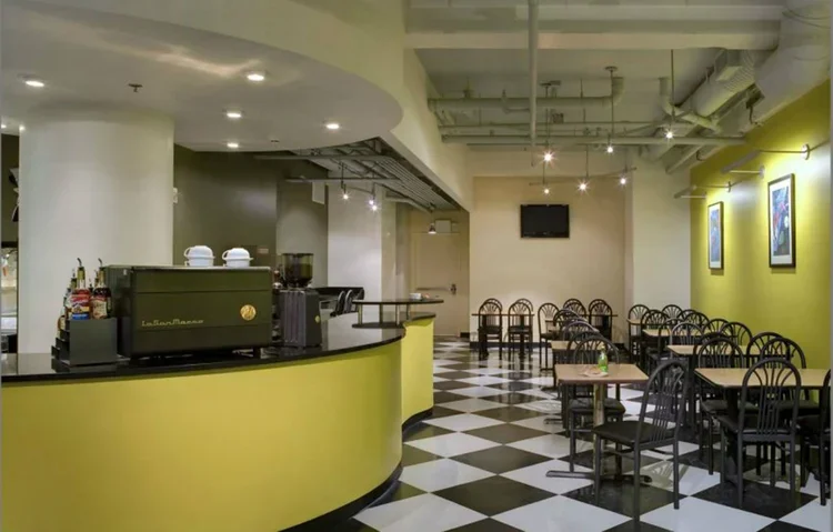 Empty cafe with checkered black and white floor, yellow curved counter, and black chairs around small tables. Wall artwork and a small wall-mounted TV are visible.